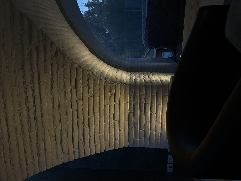 The image shows a curved wall made of white bricks, possibly part of a room or interior space. The wall has a large, rounded window cut out, offering a glimpse of trees and a building against a blue sky. A sleek, black chair sits in the foreground to the right, its back curving elegantly. The lighting is dim, with light casting shadows on the brick wall, enhancing its texture and shape.