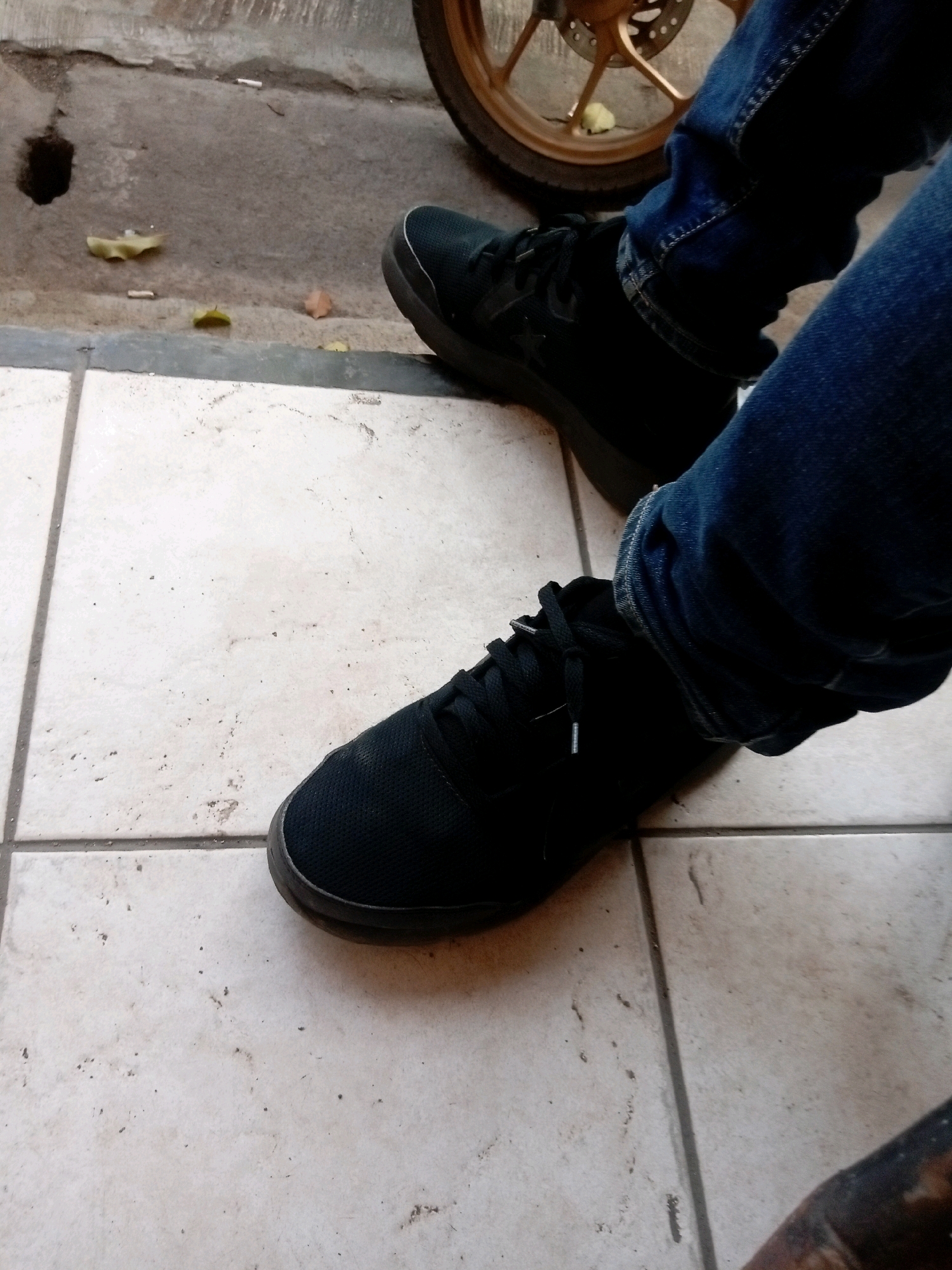 This is a close-up, low-angle, eye-level shot of a person's legs and feet, wearing dark blue denim jeans and black athletic shoes. The person is standing on a tiled floor, likely outdoors, with portions of a concrete surface visible in the background. The concrete area has scattered debris, including cigarette butts and dried leaves, and what appears to be a dark circular stain or hole. A portion of a motorcycle wheel, with a gold rim, is visible in the upper right background. The lighting suggests it is daytime. The scene appears to be in a public area, possibly near a street or walkway in Batam, Indonesia.