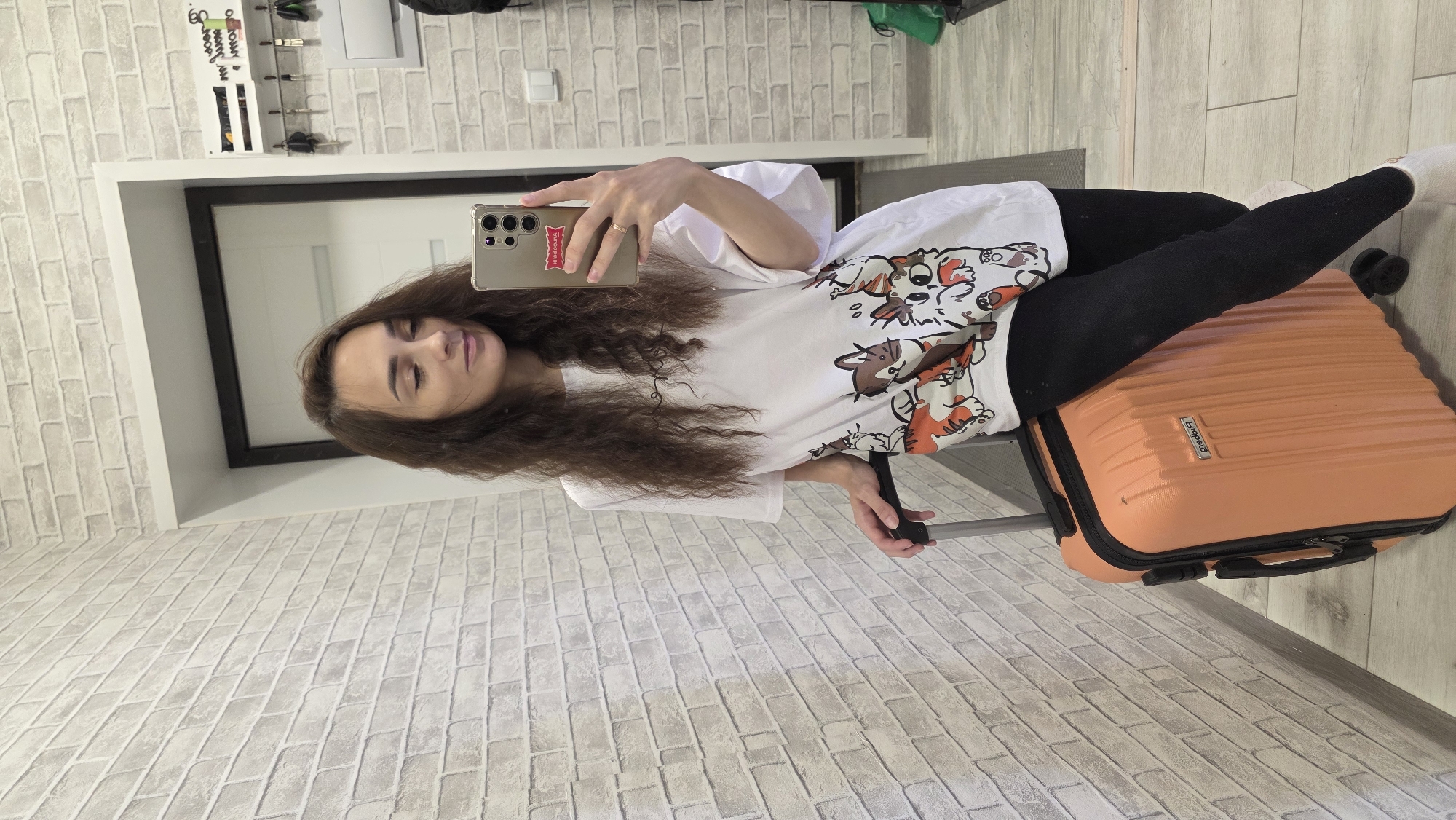 A woman with long, wavy brown hair stands in a hallway, taking a selfie with her phone. She is wearing a white t-shirt with cartoon cat prints and black leggings. To her left is an orange suitcase. The hallway has light brick-patterned wallpaper and a light-colored floor. In the background, a doorway leads to another room. A key holder with several keys is mounted on the wall to the right. The scene appears to be indoors, likely in a residential setting. The woman's phone is held up, capturing her reflection.