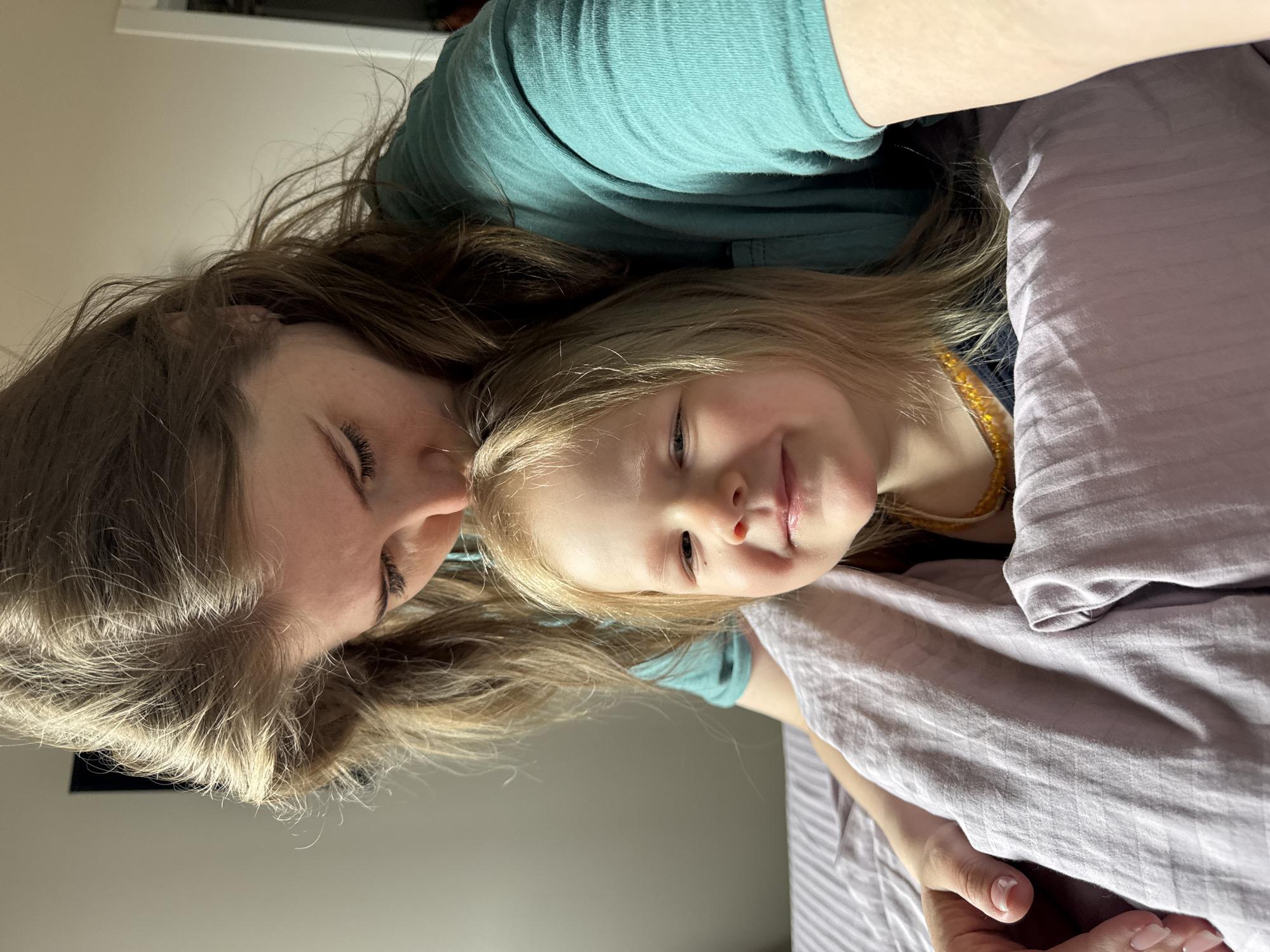 A woman and a young child are lying down close to one another. The woman has brown hair and is wearing a teal shirt. Her eyes are closed. The child has blonde hair and is wearing a yellow necklace. The child is lying on a light purple surface, likely a bed. The location is indoors and a window is visible. There is no indication this photo was taken in Ферма, Russia.