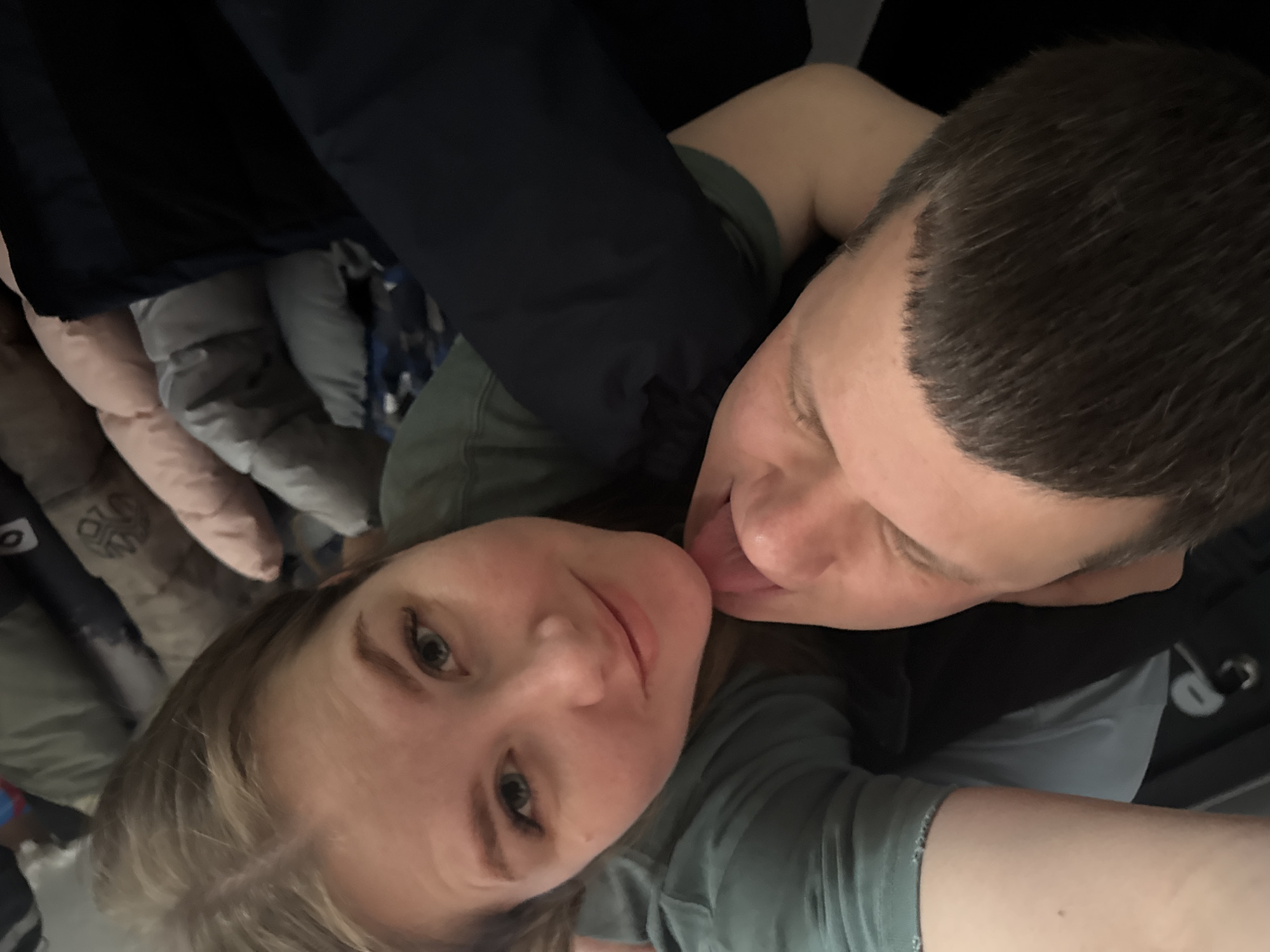The image shows two individuals taking a selfie. One is a young woman with fair skin and light brown hair, wearing a green short-sleeved shirt. She is looking at the camera with a slight smile. The second individual is a young man with fair skin and short dark hair. He is positioned behind the woman, appearing to lick her cheek. The background includes a dark jacket and a pillow with a possible pattern. The specific location within Gornyi (Dvurechenskoe S/P), Russia, cannot be determined from the image alone.