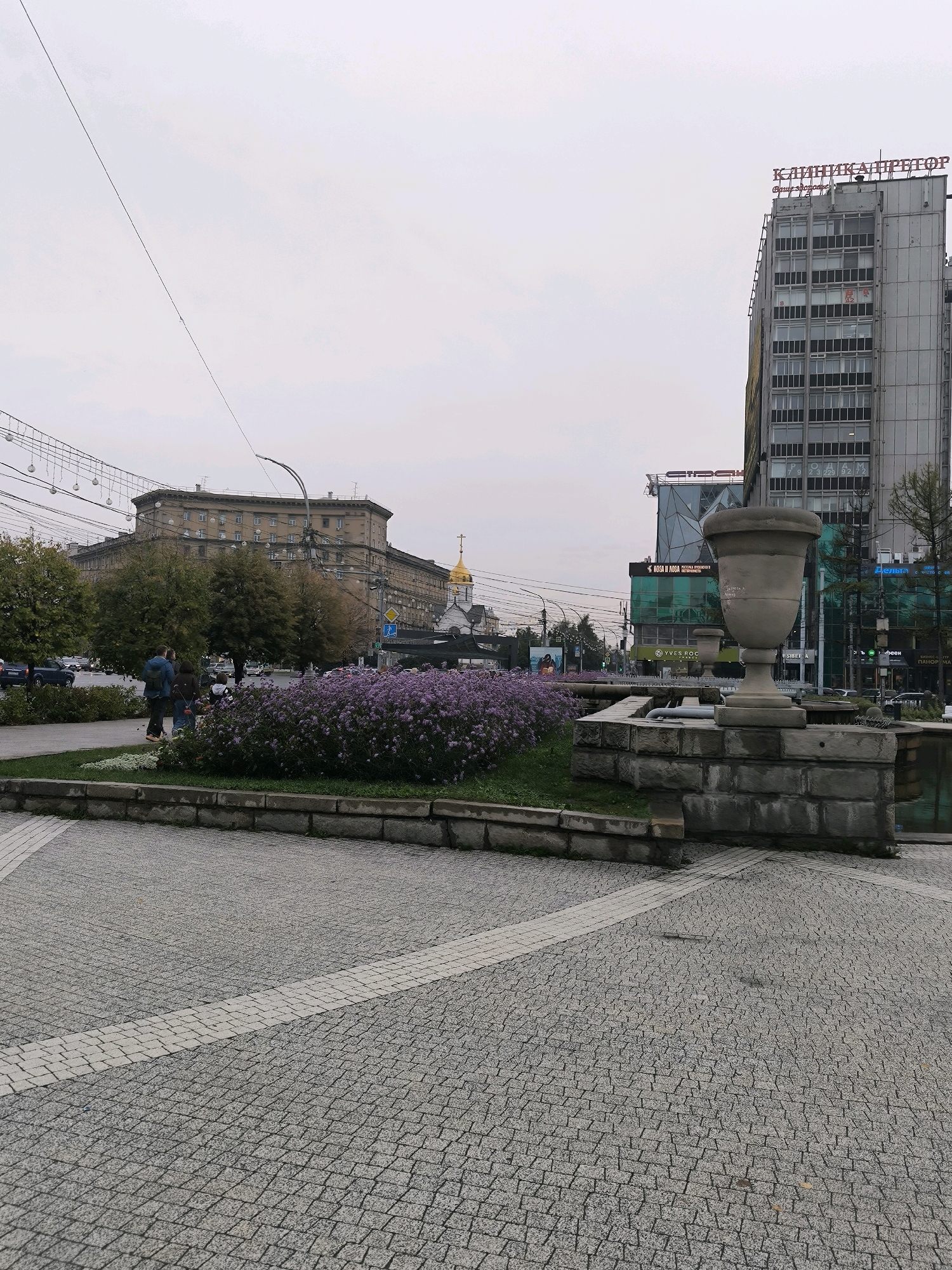 An overcast day in Novosibirsk, Russia, features a street scene with a prominent multi-story building bearing Russian text. The building's facade is largely glass and metal, with windows arranged in a grid pattern. To the left of this building, a larger, older structure with a distinctive architectural style is visible, featuring numerous windows and a curved facade. In the foreground, a paved area with a cobblestone pattern leads to a raised flower bed filled with purple flowers. A stone fountain with a large urn-like structure sits to the right of the flower bed. Several people are walking in the middle ground, near the flower bed, and cars are visible on the street in the background. Overhead, strings of lights and electrical wires crisscross the sky.