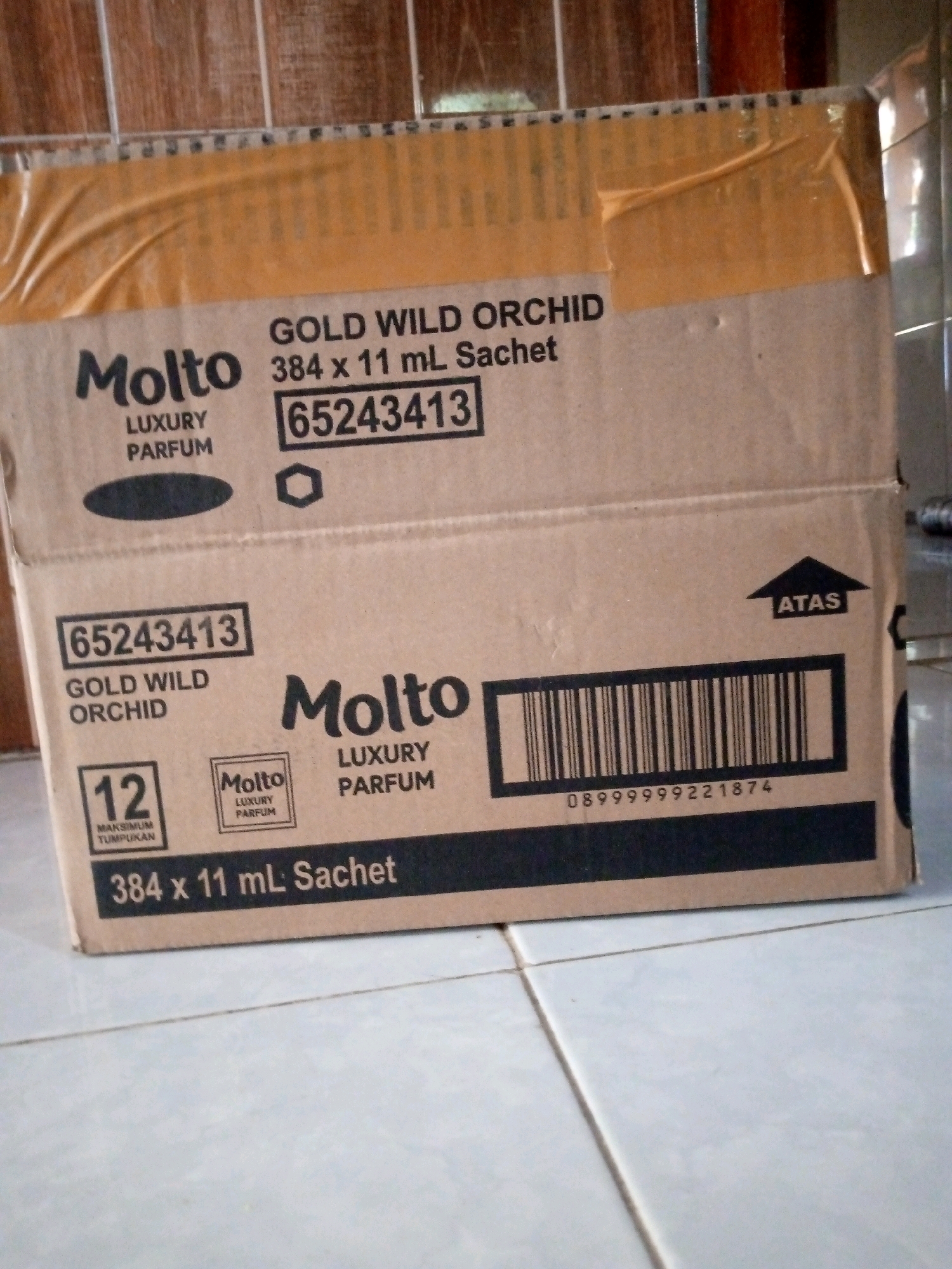The image shows a cardboard box of Molto Gold Wild Orchid laundry perfume sachets. The box is taped shut with brown packing tape and has text and barcodes printed on it. Some of the text is in Indonesian, including "LUXURY PARFUM" and "ATAS," which means "UP" and indicates the correct orientation of the box. There is also a number "12" with text below it that says "MAKSIMUM TUMPUKAN," meaning "maximum stacking." The box is placed on a tiled floor, possibly in a storage area or a warehouse. The background shows wooden planks and a tiled wall. The overall scene suggests a commercial or logistical setting in Sedati, Indonesia, where this product is likely being stored or prepared for distribution.