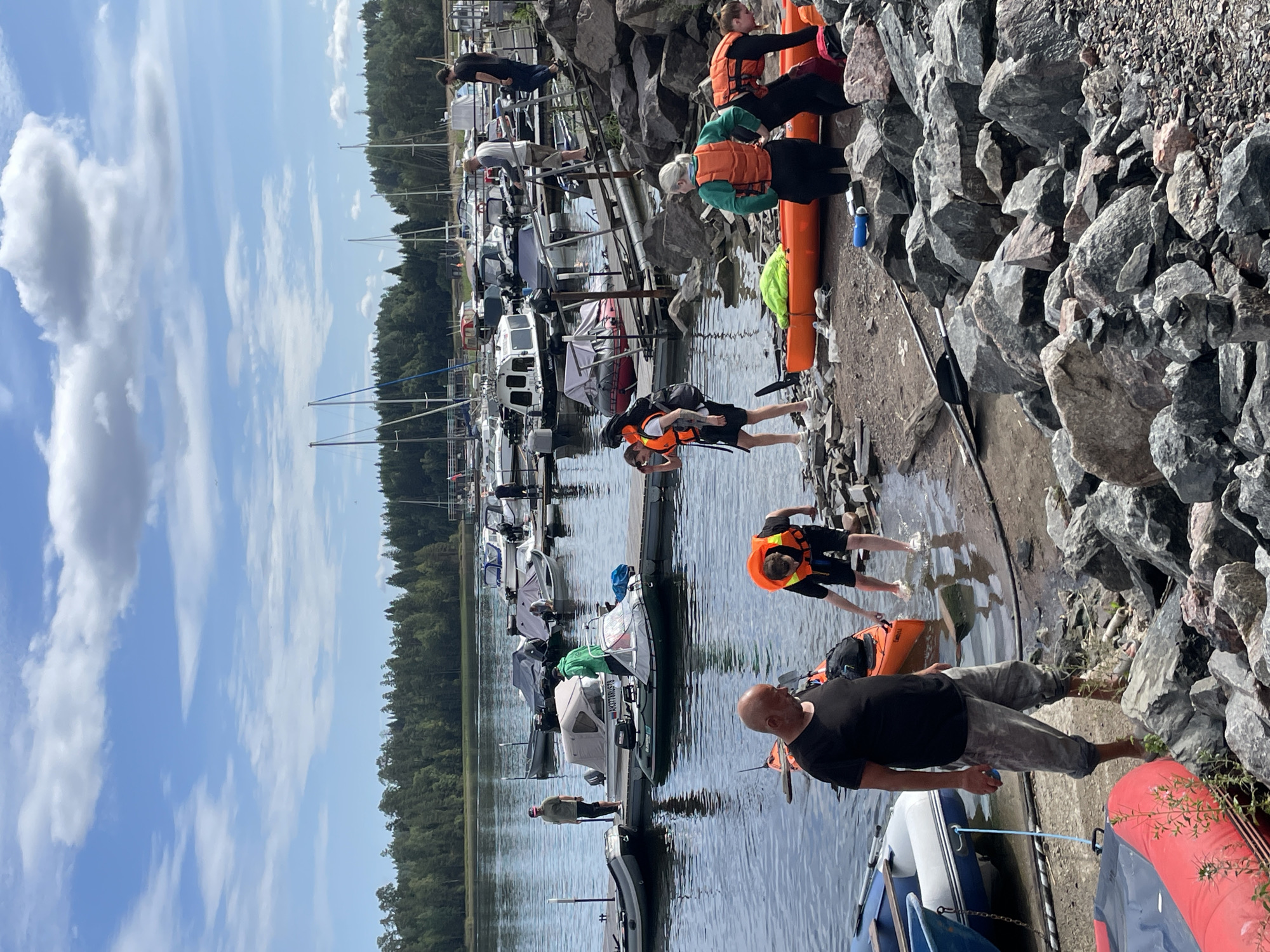 This is a full-frame, eye-level, outdoor, vertical shot taken on a cloudy but bright day at a marina. The scene depicts several people, some wearing life vests, interacting with kayaks and boats near a rocky shore. In the foreground, a man in a black t-shirt and grey pants stands facing right, with a kayak behind him. To his right, a person in a bright orange life vest bends down near the water's edge. Further back, three people are visible near an orange kayak on the shore. The water is calm, reflecting the sky and the boats docked in the background. A line of trees forms the horizon. The marina is populated with various boats of different sizes, including sailboats and smaller motorboats. The overall atmosphere suggests preparations for or conclusion of water activities.