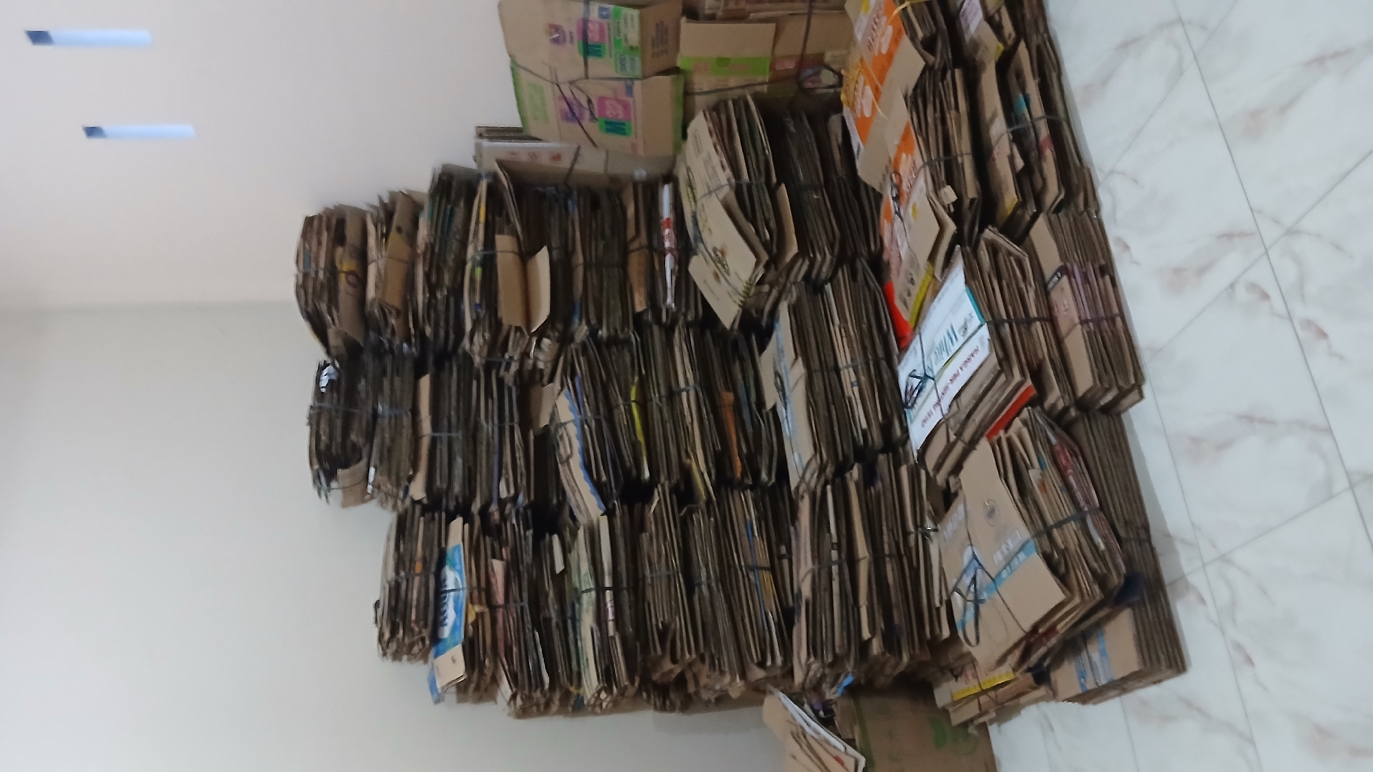 The image captures a large quantity of flattened cardboard boxes, meticulously bundled and stacked within an indoor setting. The bundles, varying in size and featuring diverse printed patterns and colors from their original packaging, are tied with strings or plastic straps. They are densely arranged, occupying the central and lower portions of the frame, resting against a plain white wall.
The scene depicts an organized collection or storage area, likely for recycling or resale. A light-colored, marble-patterned tiled floor is visible on the right side of the image. The room's illumination suggests either artificial light or indirect natural light, indicating an indeterminate time of day.
Among the visible text on some of the bundled cardboard, a clear phrase reads "W/HARGA TER" and "HARGA PER SATUAN/PER BUNGKUS" (price per unit/per bundle), indicating pricing or inventory information. Other faint texts and graphics are present on various boxes but are not clearly legible. The overall impression is one of systematic waste management or material handling in Lhokseumawe, Indonesia. The image itself appears to be rotated 90 degrees counter-clockwise.