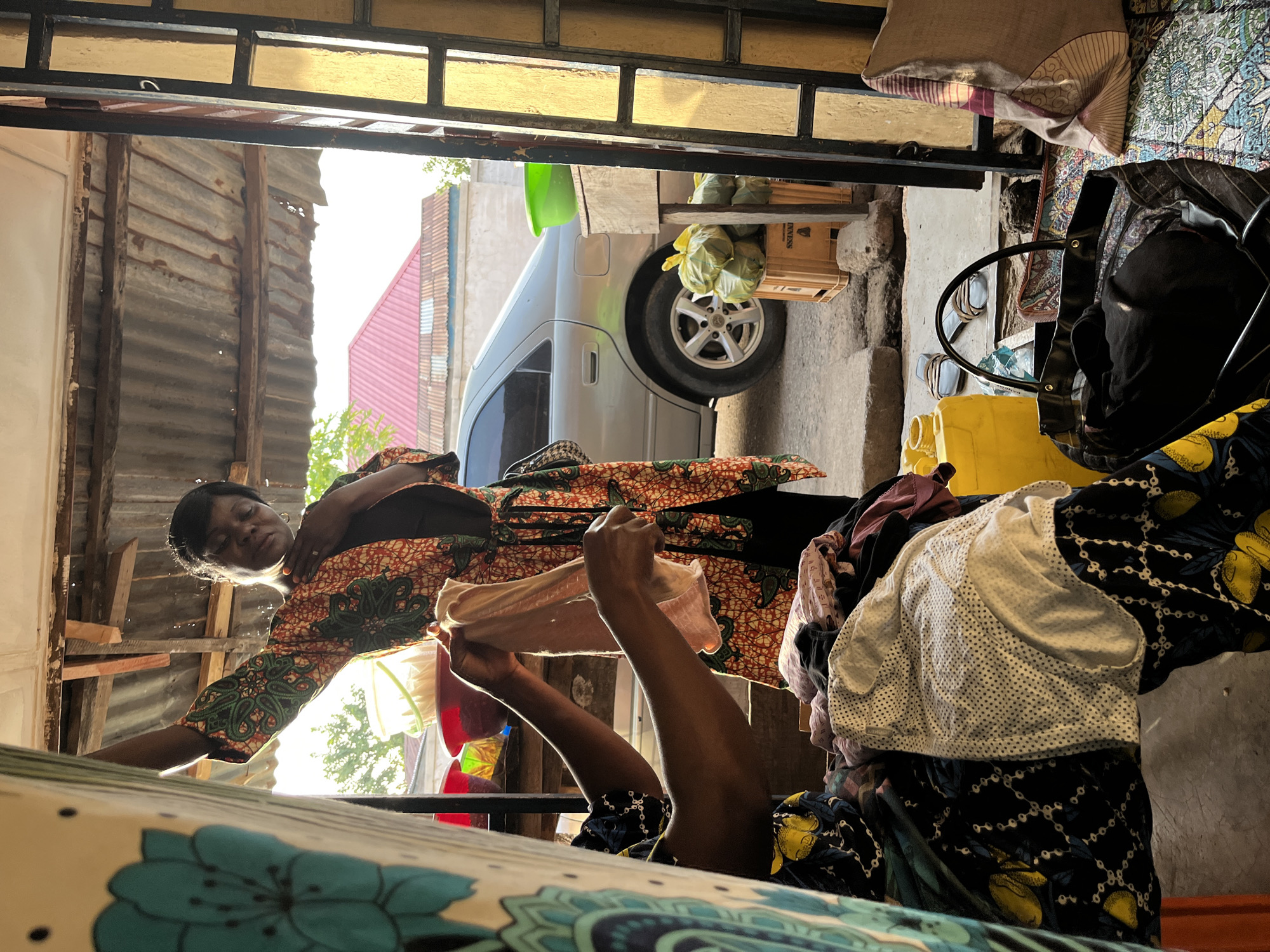 The image depicts a woman inside a structure with corrugated metal walls, possibly a shop or home in Jos, Nigeria. She is wearing a patterned dress and holding up a garment to examine it. The foreground shows a patterned cloth draped over a surface. Through the open doorway, a vehicle is visible outside, along with other items such as a wooden crate, a yellow container, and a black bag on a chair.