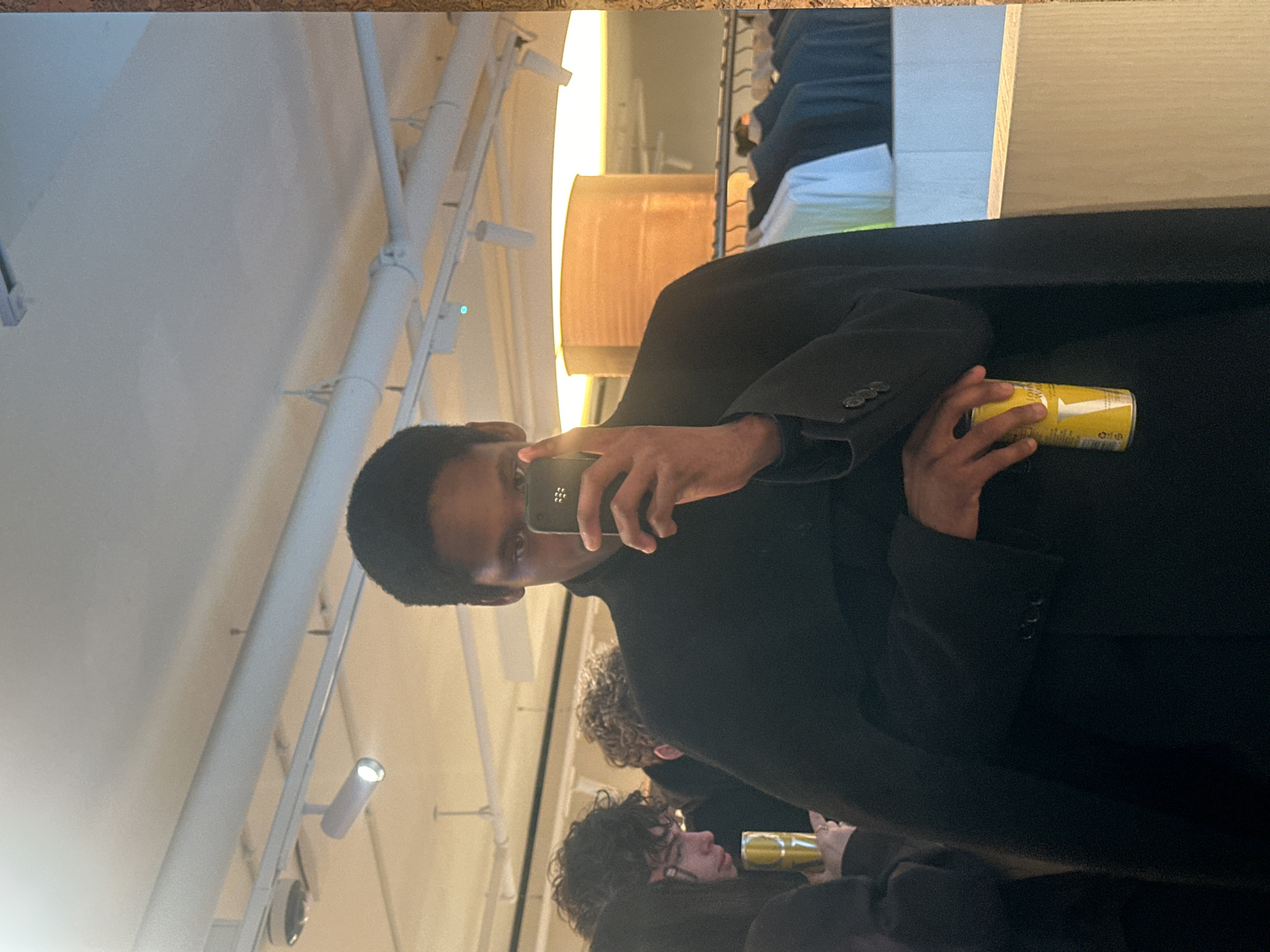 An interior mirror selfie captures a modern retail or event space in the city of London, United Kingdom.
A young adult male, appearing of Black ethnicity, with short dark hair, stands centrally, visible from the chest up. He holds a dark-colored smartphone in his right hand, partially obscuring his face while taking the photograph. His dark eyes are visible above the phone. He wears a dark, high-neck garment, possibly a turtleneck, beneath a dark, broad-shouldered cape-like or oversized coat/blazer. In his left hand, positioned below his right arm, he holds a yellow cylindrical beverage can with white text and a prominent white "V" logo, indicative of a Johnnie Walker Highball.
To the subject's left, a person with dark, curly hair and spectacles is partially visible, holding a translucent yellow cup. Behind this individual, another person with curly hair is partially obscured.
The reflected background features a white ceiling with exposed light grey pipes, track lighting fixtures, and a small green indicator light. A brightly lit spotlight is visible on the track. A large cylindrical light fixture emits a warm orange glow in the mid-right background. To the right, a clothing rack displays various garments, including light blue/green and darker items. A light-colored wooden or laminate display surface is visible in the lower right, with a light blue-grey wall section behind the clothing rack. The mirror has a visible light-colored, possibly corkboard or textured wood, frame on its right edge. The overall environment suggests a contemporary commercial or event setting with industrial-chic design elements.