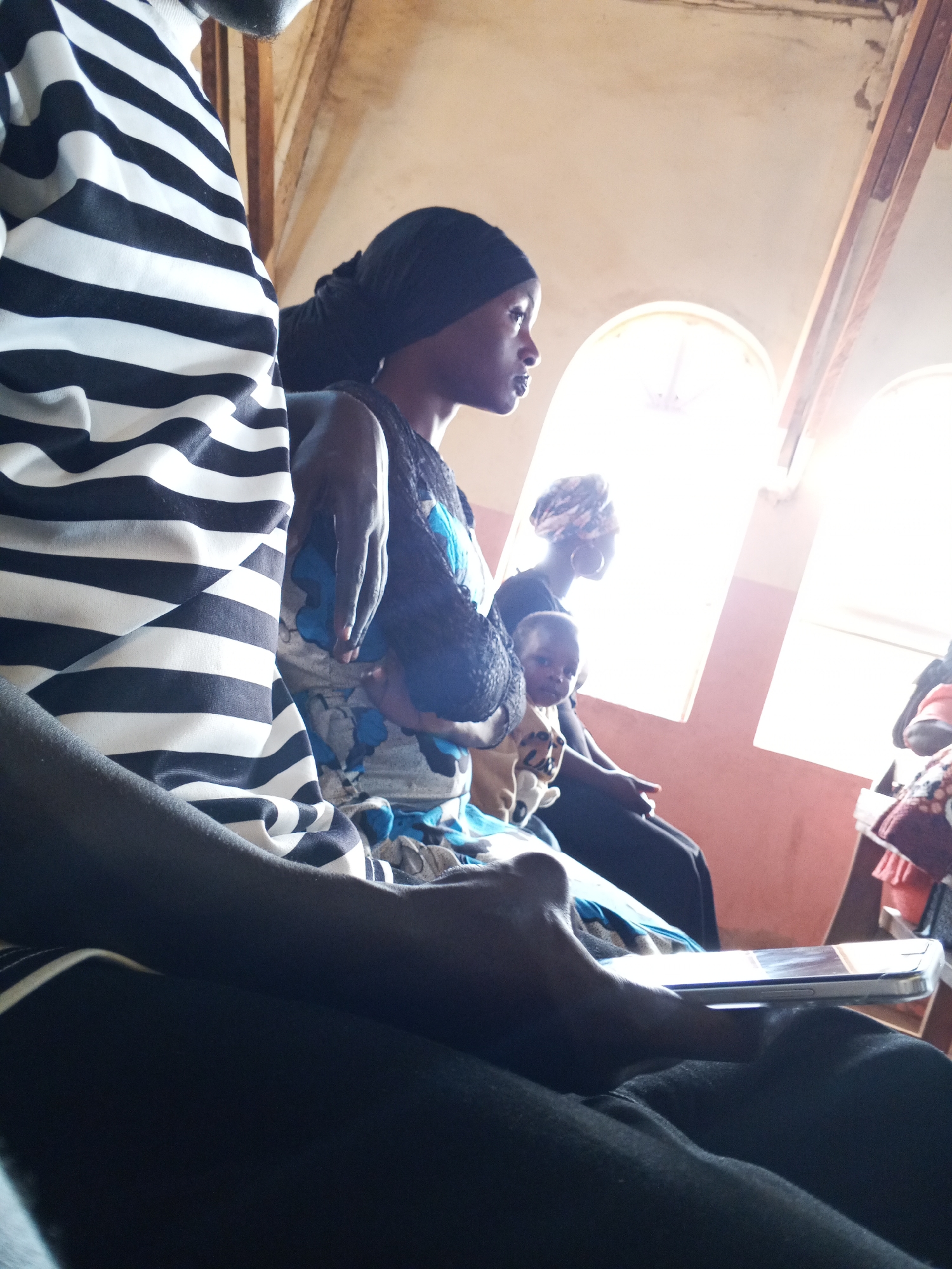 The image shows a group of people inside a building, likely a church, in an unknown location. A woman with a head covering is holding a baby, and another woman is seated behind them. In the foreground, a person wearing a black and white striped shirt is holding a smartphone. The building has arched windows and a peach-colored wall.