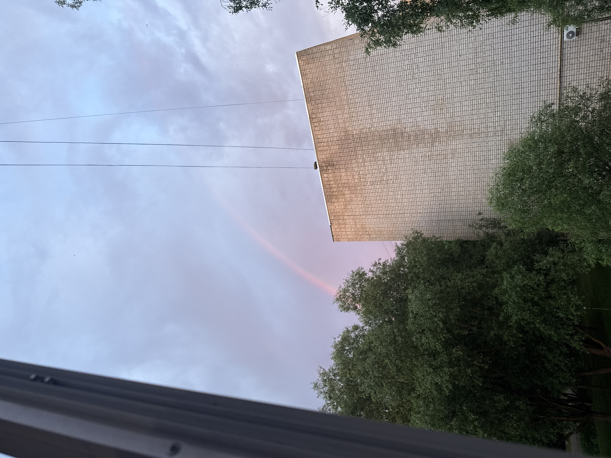 A rainbow is visible in the sky over a residential building in Valday, Russia. The building is made of light-colored bricks. Tall trees with green leaves are in the foreground, obscuring the lower part of the building and the ground. The sky is cloudy with hues of pink and blue, suggesting either sunrise or sunset. Electrical wires are suspended in the air. The view appears to be from a window.