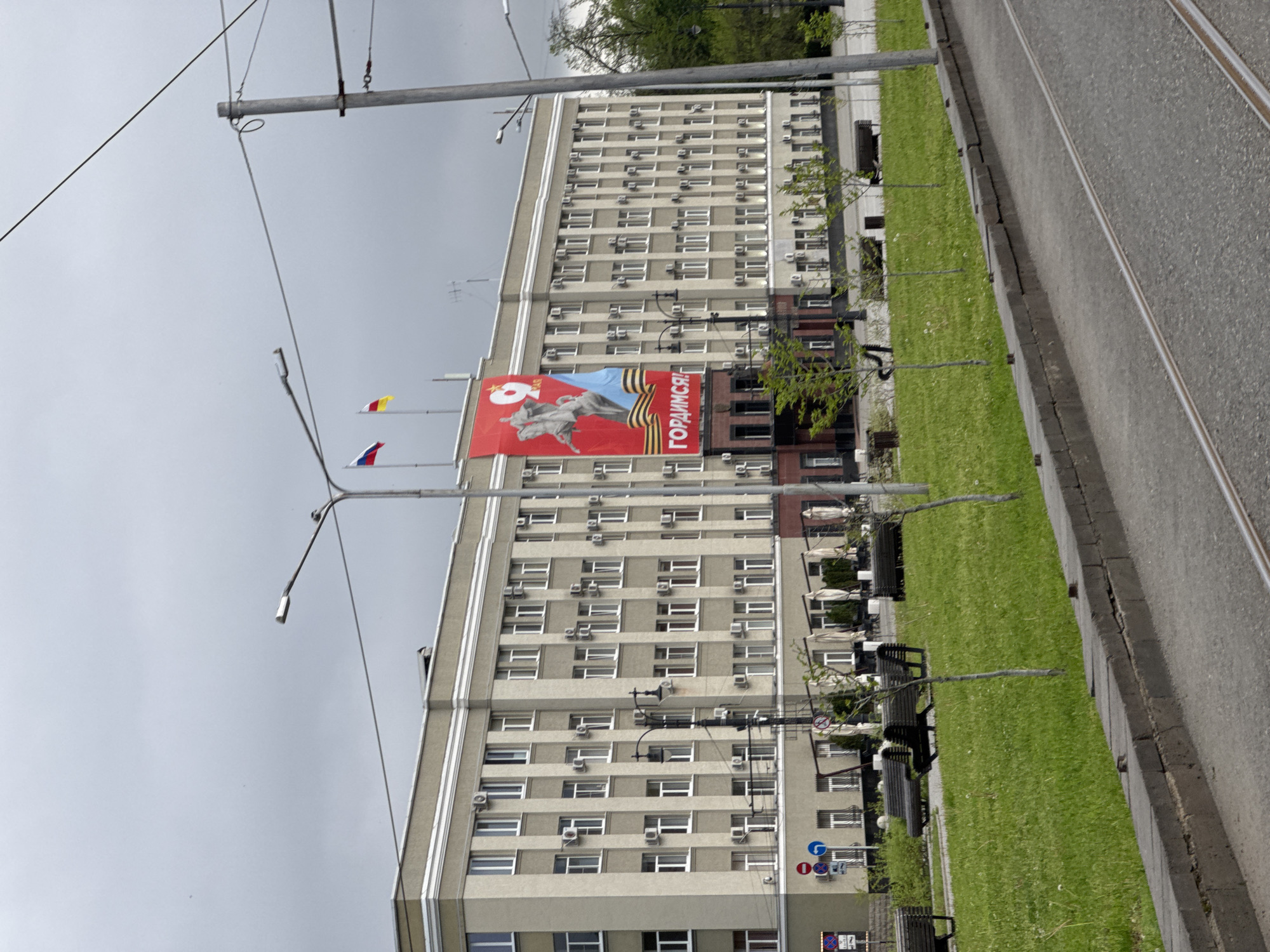 A vertical shot shows a tall building with many windows and air conditioning units, situated beside a patch of green grass and a street with tram tracks. A large red banner with Russian text and imagery of a statue hangs on the building. There are two flags atop a pole. The sky is overcast. The location is unknown.