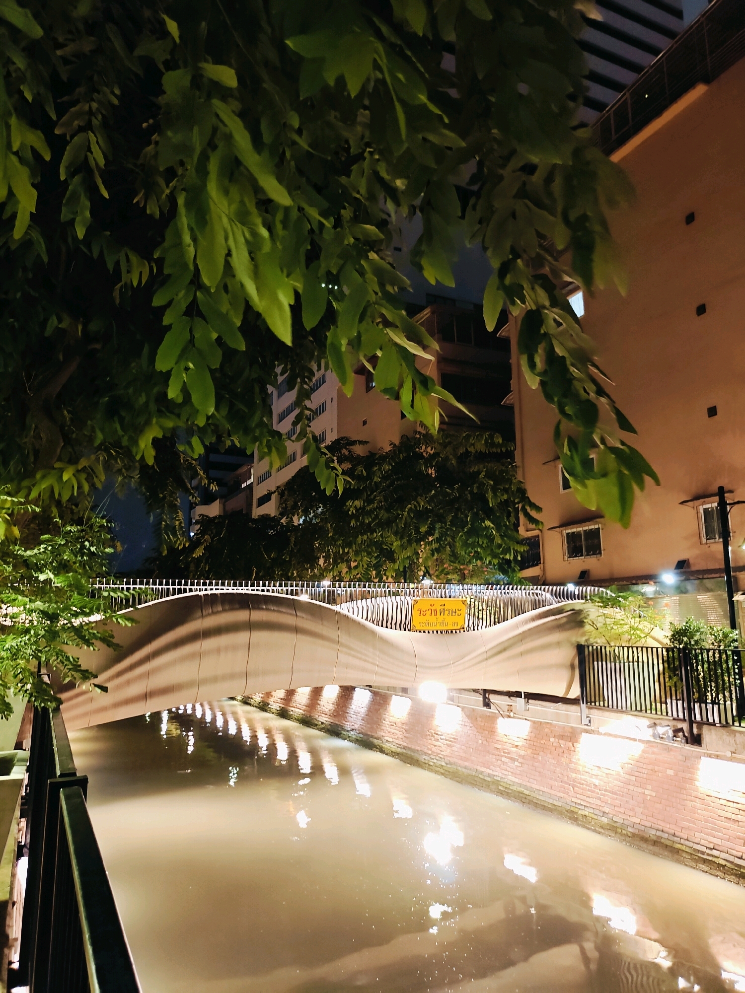 This is a nighttime, low-angle photograph of a pedestrian bridge over a canal in Bangkok, Thailand. The bridge has a modern, curved design with a light-colored, flowing surface. Above the bridge, the leaves of a tree hang down, partially obscuring the view of the buildings behind. The canal water is murky and reflects the lights from the bridge and the surrounding buildings, which include a brick-lined canal wall with lights casting reflections. A yellow sign with Thai text is visible on the bridge's railing. The scene appears to be in an urban environment, given the presence of buildings and the canal.