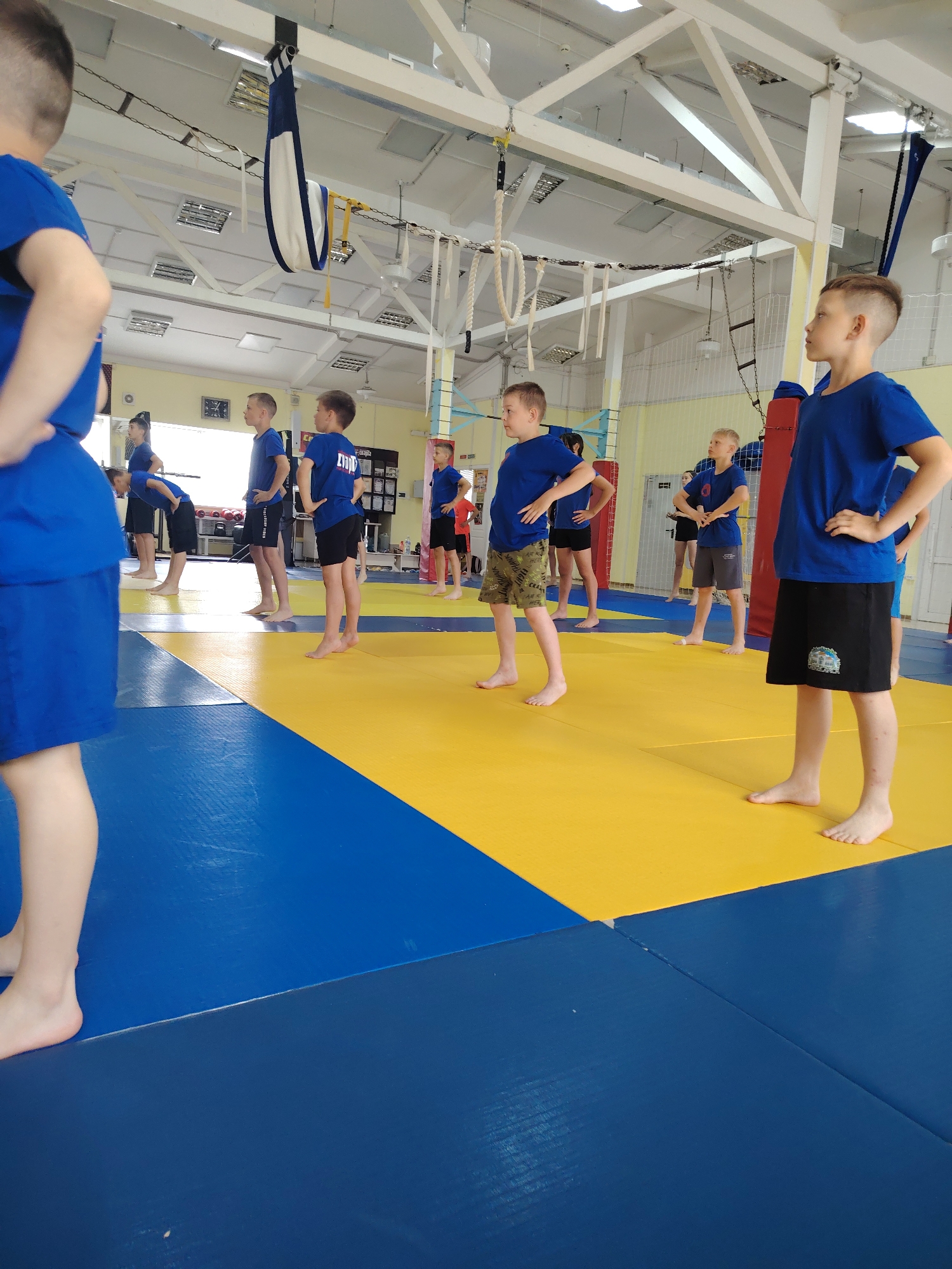 The image displays a group of young boys engaged in a training session within an indoor gym or sports hall in Chaikovsky, Russia. The setting features a spacious room with a high white ceiling supported by metal trusses and illuminated by fluorescent lights. The floor is covered with distinctive interlocking mats in vibrant blue and yellow, typically used for martial arts or physical education.
The subjects are pre-teen boys, predominantly dressed in blue t-shirts and various types of shorts, including black, camouflage, and grey. All are barefoot, standing in rows with their hands on their hips, indicating they are likely performing warm-up exercises, stretching, or listening to instructions. Their expressions are generally neutral and focused.
In the background, gym equipment such as hanging straps, ropes, and red padded columns are visible, along with white walls and a clock. The ambient light suggests it is daytime.
Visible text includes "LIVE FOR THE HILLS" on the black shorts of one boy, and a partially obscured word, possibly "ZYDOP", on the blue t-shirt of another. A small, indistinct white logo can also be seen on a different pair of black shorts. The scene captures a structured physical activity session for children.