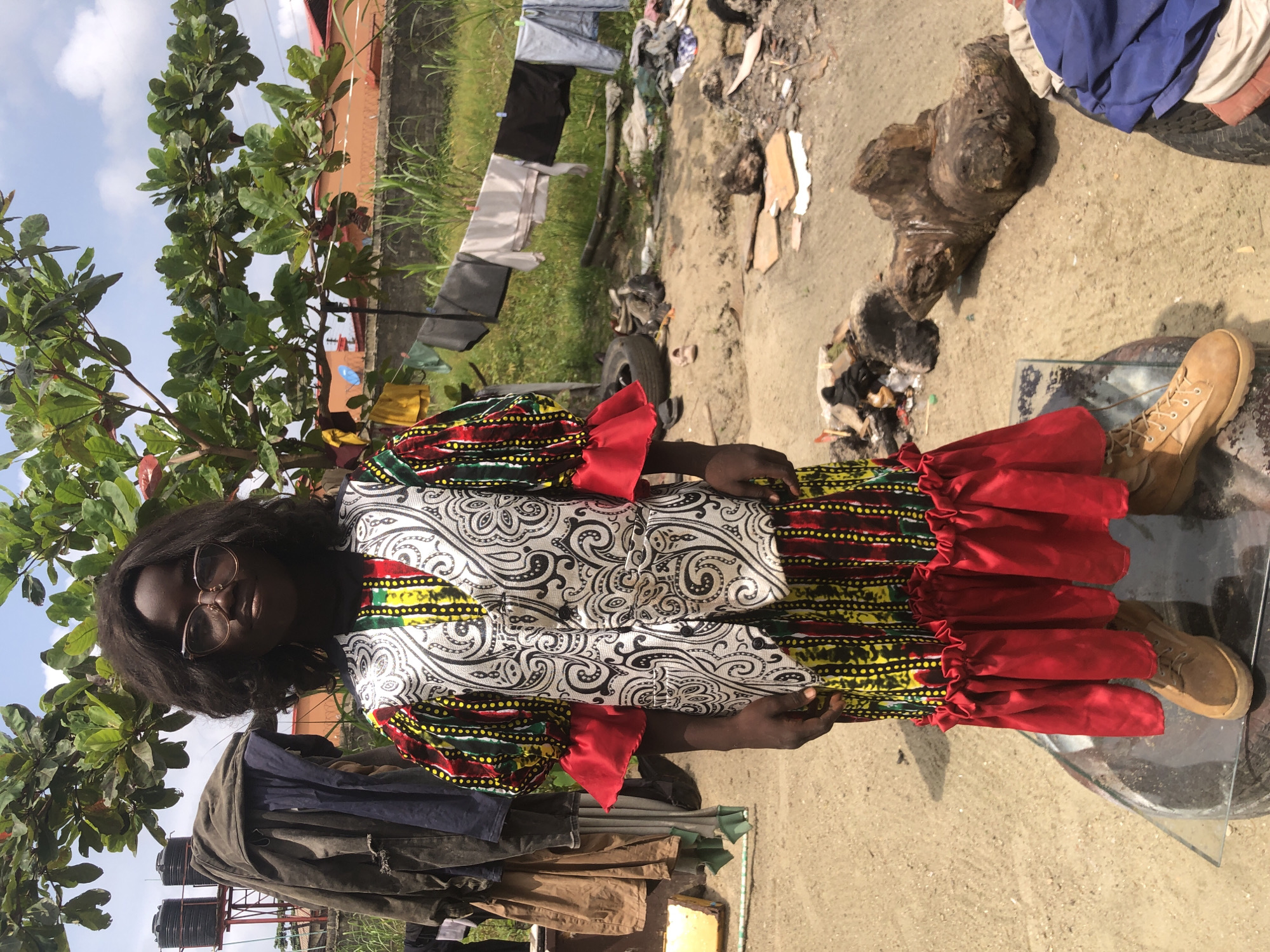The image shows a young girl standing in what appears to be an outdoor, possibly informal, settlement. The setting is characterized by a dusty ground, scattered debris, and a clothesline with laundry hanging in the background. A tree with green leaves provides shade overhead. The girl is wearing a vibrant, colorful traditional African outfit, consisting of a patterned dress with a vest over it. She has on glasses and tan boots. The environment suggests a warm climate. The presence of water tanks and some basic structures in the background might indicate a residential area within Ado, Nigeria. The overall scene depicts everyday life in a community, likely in a developing area.