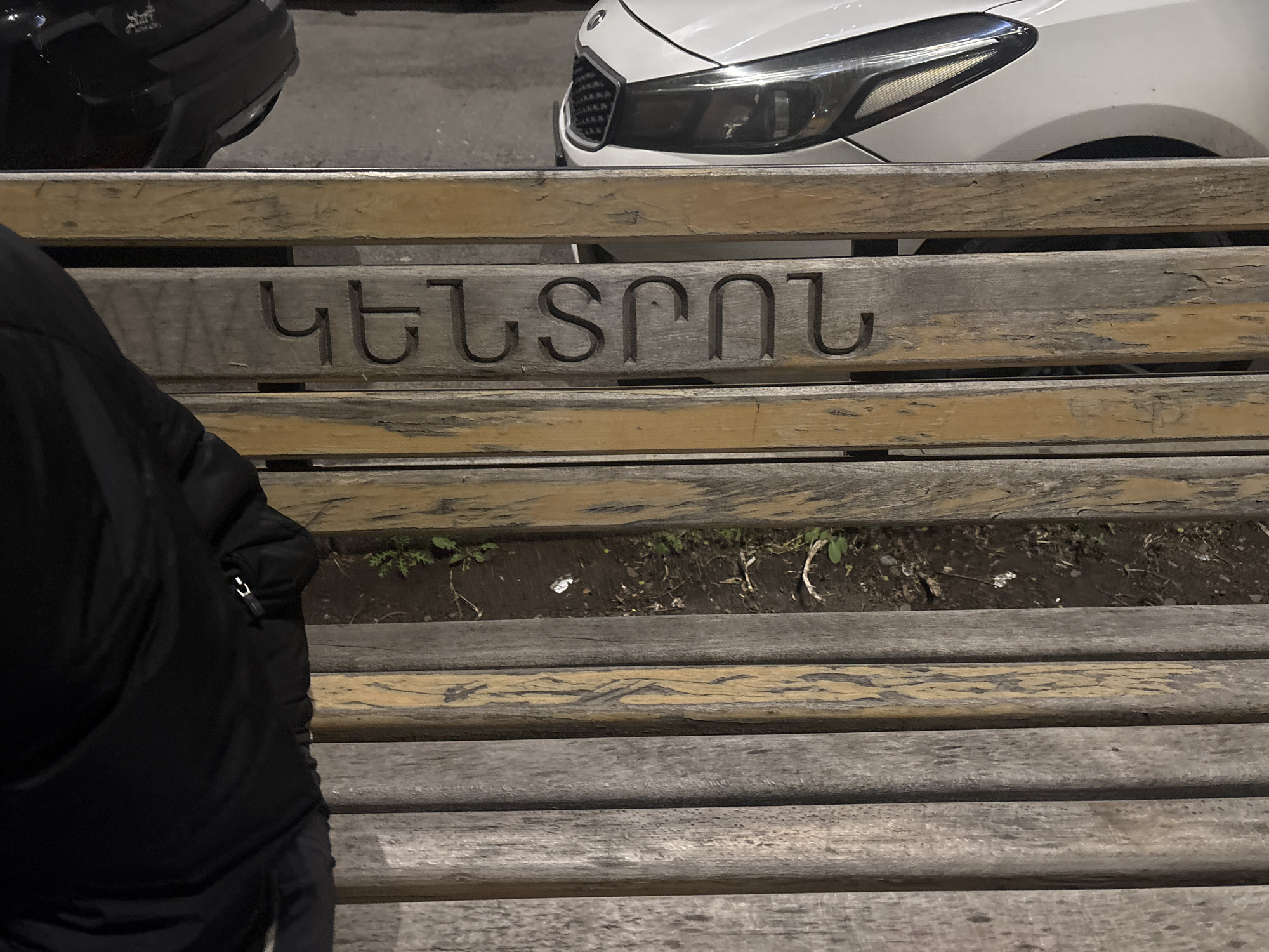 The image shows a weathered wooden bench with the word "ԿԵՆՏՐՈՆ" carved into it, which translates to "Center" in Armenian. A black jacket is visible on the left side of the bench, partially obscuring it. Behind the bench, the front of a white Kia car is visible, along with the back of a black car. The scene is likely in a public area, given the bench and the presence of vehicles, and is inferred to be in Yerevan, Armenia due to the Armenian word carved into the bench.