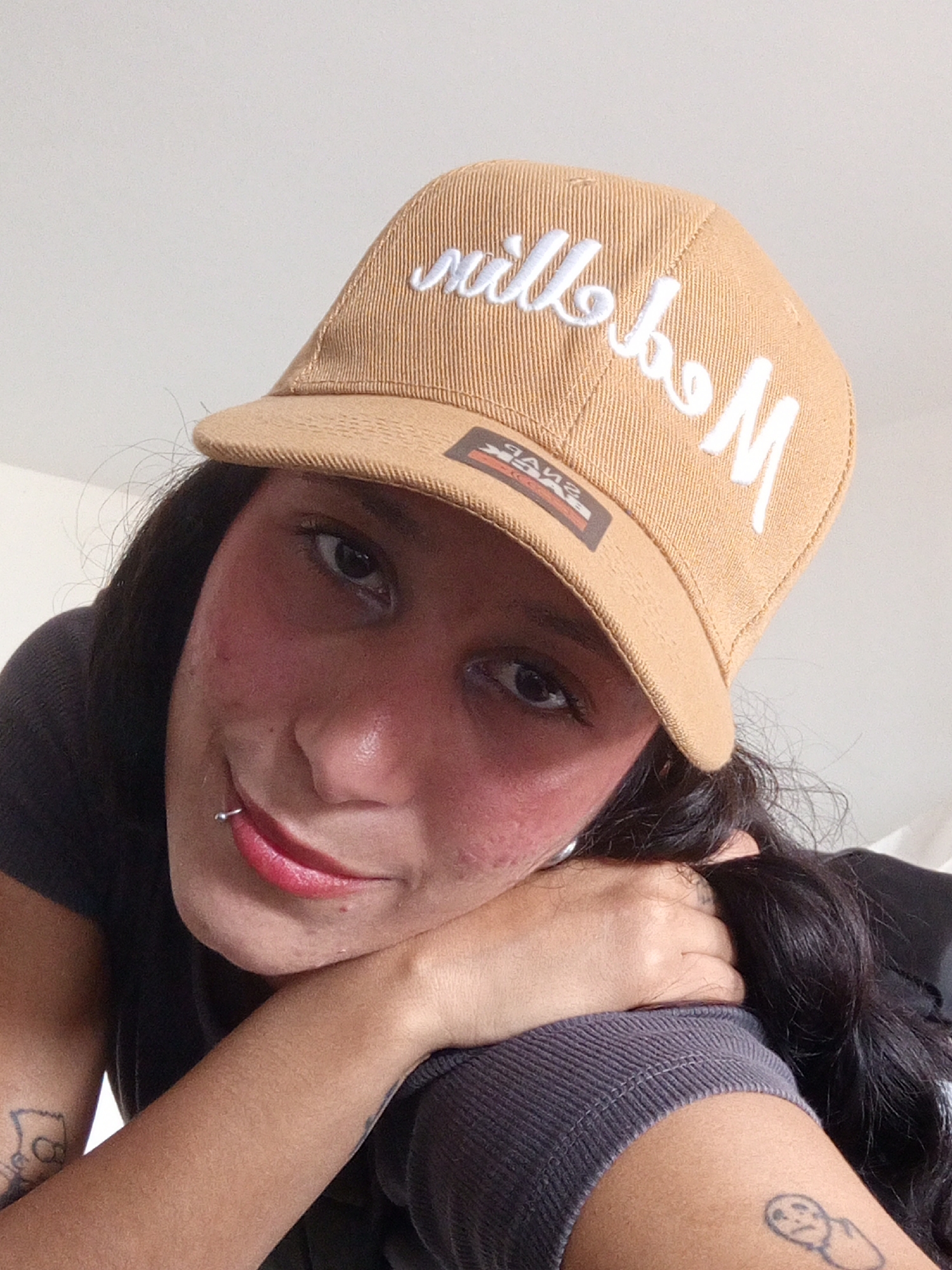 A close-up, upward-angle shot of a light-skinned person wearing a tan baseball cap with white embroidered lettering that appears to read "Millboy M." The person is leaning their head on their arm, which is clad in a gray long-sleeved shirt. The person has dark hair and is wearing red lipstick. A small metal lip ring is visible. There are tattoos visible on both arms, one in black ink near the shoulder and another on the forearm. The background is a blurred white wall, likely indoors. There is no information in the provided image to determine the location.