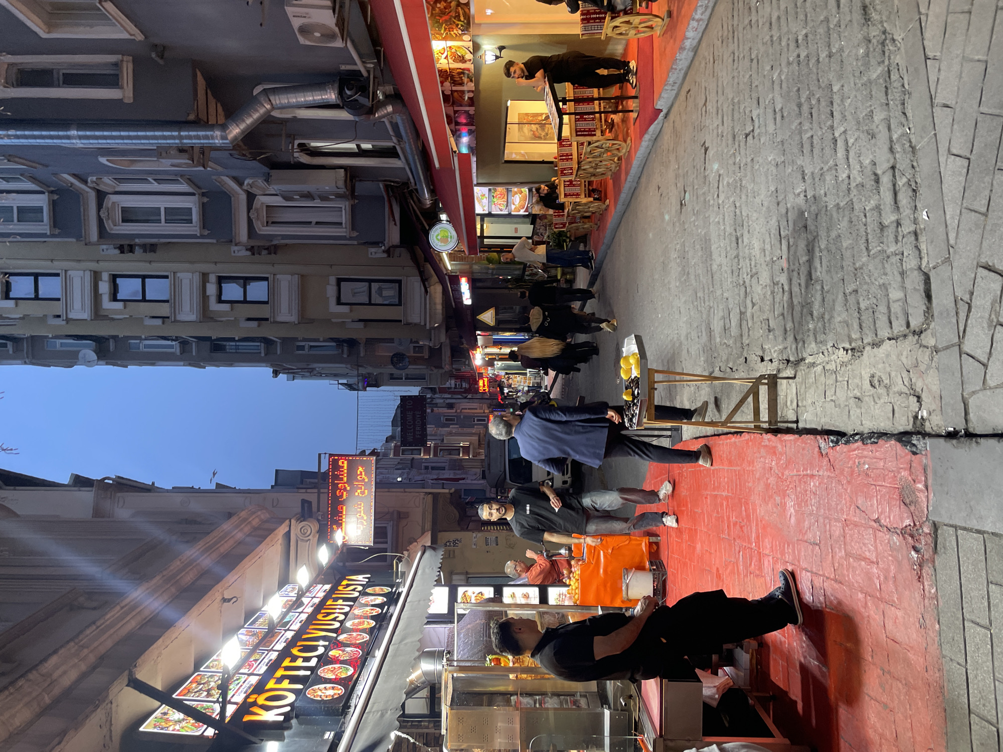 The image captures a street scene in Beyoğlu, Turkey. The view looks down a narrow street, with buildings on either side. On the left is a restaurant with an illuminated menu board featuring the name "KOFTECI YUSUFUSTA" and an additional sign with Arabic script. Adjacent to the restaurant, along the left side of the street, is a red-painted sidewalk section where individuals are seated and standing. Some are engaged in conversation. On the right side of the street, a similar red-painted sidewalk extends outwards, with a person standing and leaning over a menu board. Further down the street, people are walking. The street itself is paved with gray stones. The sky is a clear dusk blue. The buildings are multiple stories, with windows and architectural details. There are streetlights and overhead wires.