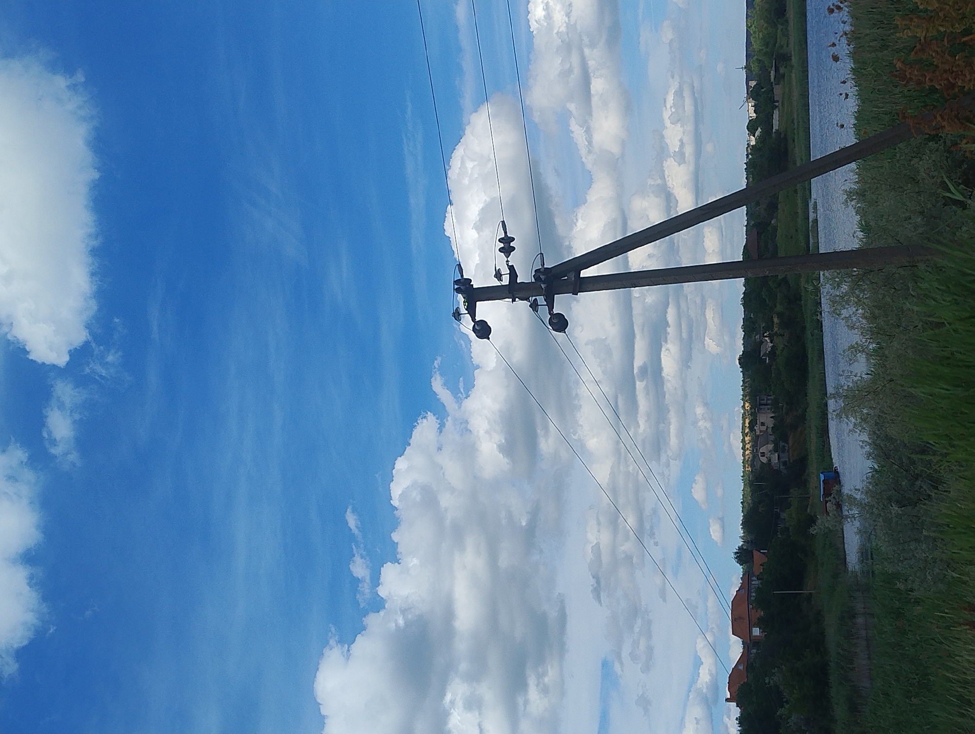 A bright, sunny day in an outdoor setting with a clear blue sky, scattered white clouds, and a body of water. In the foreground, a wooden utility pole with wires is prominent, supporting electrical transmission lines that stretch across the frame. Beyond the pole, a river or lake is visible, bordered by dense green reeds and vegetation. In the distance, a line of trees runs along the bank, with houses and buildings peeking through the foliage. The scene suggests a rural or suburban environment, possibly near a waterway.