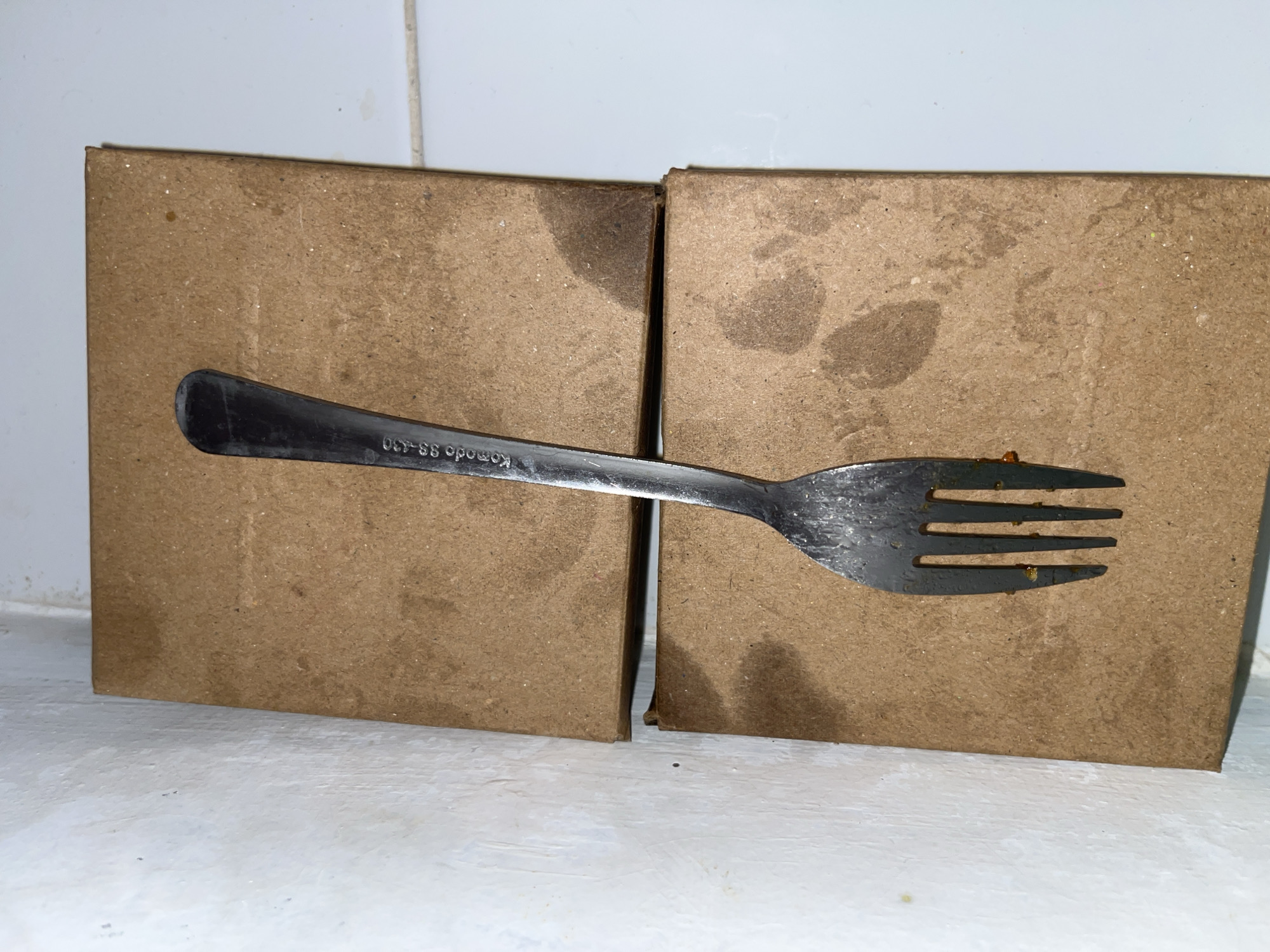 This image, taken in Lhokseumawe, Indonesia, depicts a used metallic four-tined fork resting vertically on two stacked, rectangular brown objects. The fork, positioned slightly left of center, displays an inscription "Komodo SS-430" on its handle and exhibits dark residue on its tines, consistent with food remnants. The two stacked objects, possibly made of pressed wood or thick cardboard, have a rough, speckled texture and show irregular dark stains on their surfaces; a narrow gap is visible between them. To the left of these objects is a textured, off-white wall with small dark specks. To the right, a smooth white surface, likely tiled, is visible, with a faint horizontal grout line in the lower right. The scene appears to be indoors, under artificial lighting.