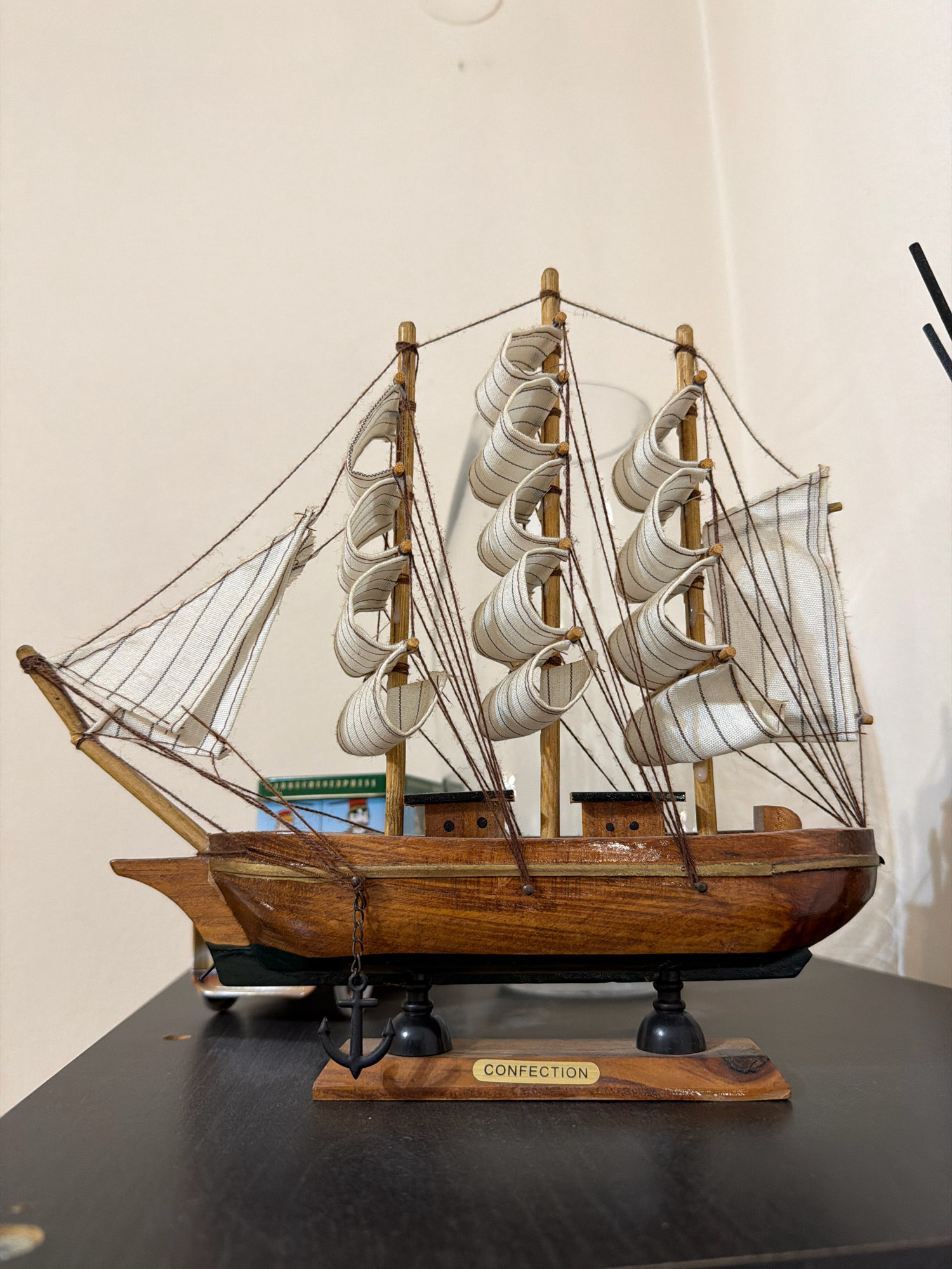 A wooden model sailboat, named "Confection," is displayed on a dark surface. The sailboat has multiple masts with white sails adorned with thin black stripes. An anchor hangs from the side of the boat, suspended by a chain. The background is a plain, light-colored wall. The model is positioned in a home or shop, and no specific location in the city of Nafplio, Greece, can be identified.