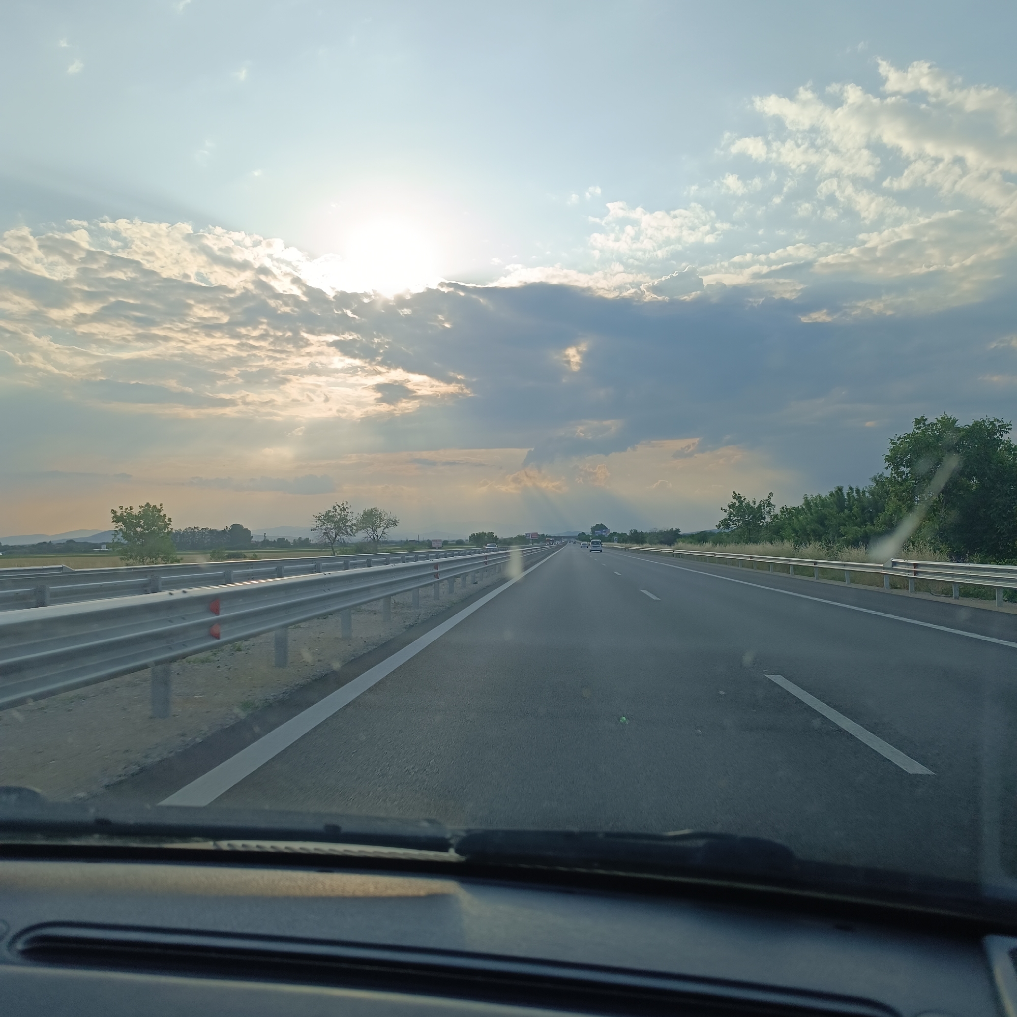 The scene depicts a multi-lane highway viewed from inside a moving vehicle, located in the vicinity of Saraia, Bulgaria. The sky occupies the upper two-thirds of the image, featuring a low, bright sun positioned in the upper-left quadrant, partially obscured by numerous clouds. Crepuscular rays (sunbeams) fan out prominently from behind the clouds, extending across the sky towards the right. The clouds vary in formation, from scattered cumulus-like structures to more continuous, darker strata.
The highway extends into the distant horizon, marked by clear white lane lines. Several light-colored vehicles are visible ahead, traveling in the same direction. Metal guardrails run parallel to the road on both the left and right sides. Beyond the guardrails, the landscape includes green trees, some appearing as clusters, and sections of lighter-colored vegetation or fields. Distant hills or mountains are faintly visible on the horizon line.
The bottom portion of the image shows the dark dashboard and the base of a windshield wiper, indicating the perspective is from the interior of the vehicle, likely the driver's or front passenger's seat. A subtle reflection or glare is visible on the windshield in the lower-right area. No individuals are directly visible in the image.