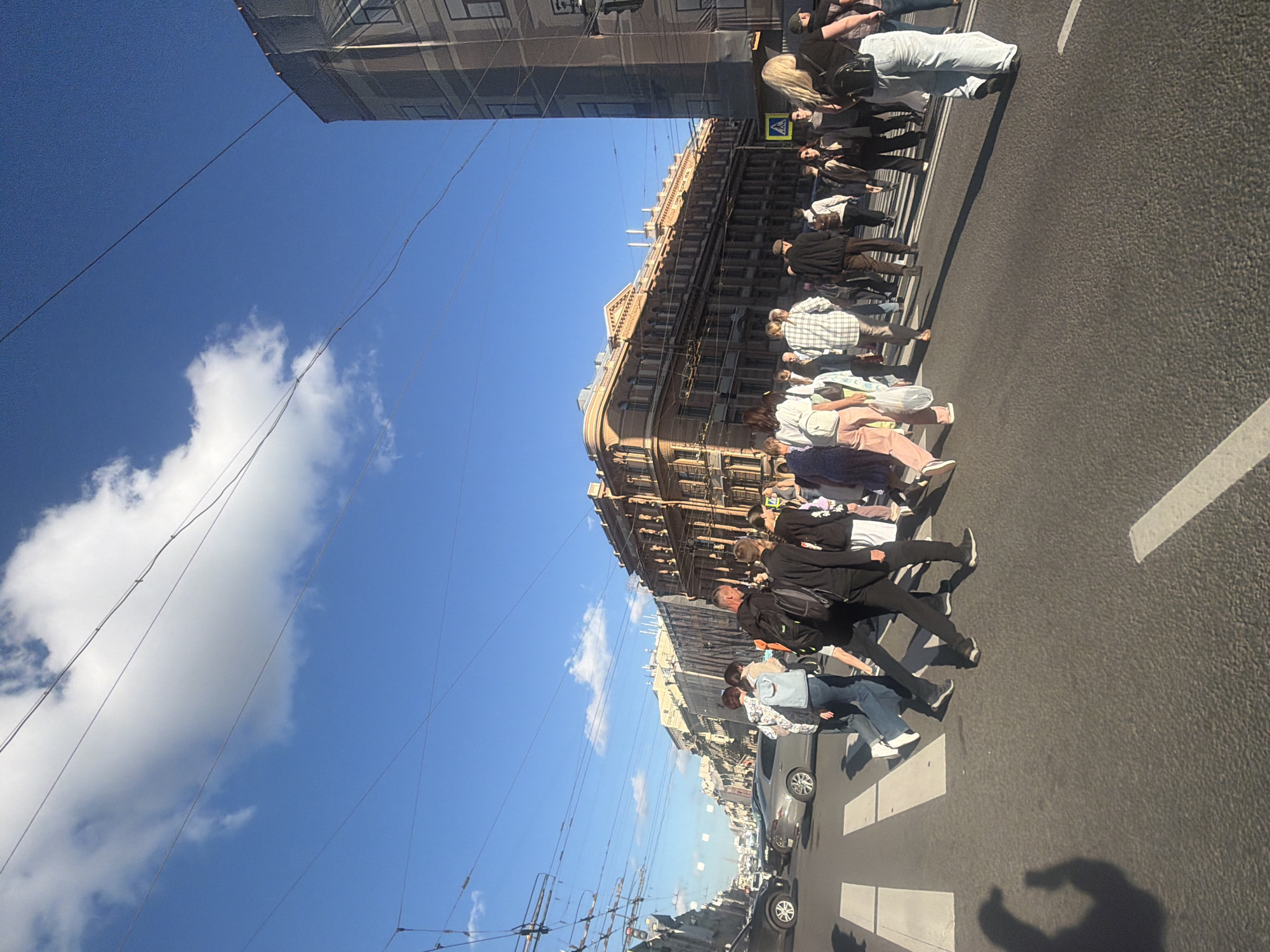 The image depicts a street scene in Saint Petersburg, Russia, featuring a pedestrian crossing on a sunny day. A diverse group of people, including men, women, and possibly adolescents, are crossing the street. Some are walking in groups, while others appear to be alone. They are dressed in casual attire, with varying colors and styles of clothing. Several individuals are carrying backpacks or bags.
The street is paved with asphalt and marked with white pedestrian crossing lines. Cars are visible on the left side of the image, stopped or moving slowly, indicating traffic. The background is dominated by the facades of old, multi-story buildings with architectural details characteristic of Saint Petersburg. Some of these buildings appear to be under renovation, as suggested by scaffolding or netting. The sky is blue with scattered white clouds. Power lines are strung across the sky, crossing over the street. The overall impression is a typical urban environment, bustling with pedestrian and vehicular activity under a bright sky.