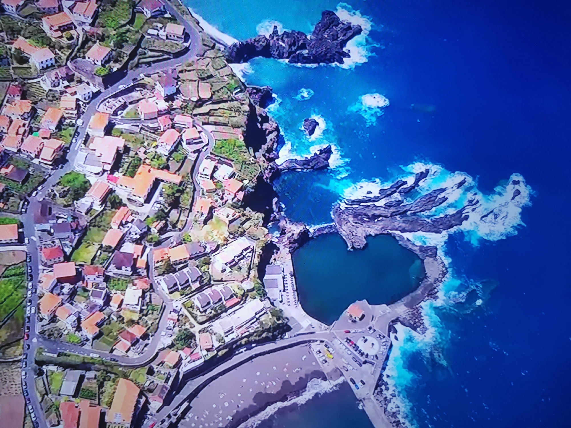 This is an aerial photograph of a coastal town with houses situated on a hillside overlooking a rocky coastline and the ocean. The buildings are predominantly white or light-colored with orange or red roofs, and they are nestled amidst green vegetation. A winding road snakes through the town, and there are several parked cars visible. The coastline is characterized by dark, jagged rocks that are partially submerged in the bright blue ocean. Waves crash against the rocks, creating white foam. There is a small, dark pool of water enclosed by the rocks, which appears to be a natural swimming area. A paved area with parked cars and a small building is located next to the pool. In the distance, the ocean extends to the horizon. There are no visible people in the image, and the scene appears to be a quiet, scenic coastal village. This image is not located in Dnipro, Ukraine.