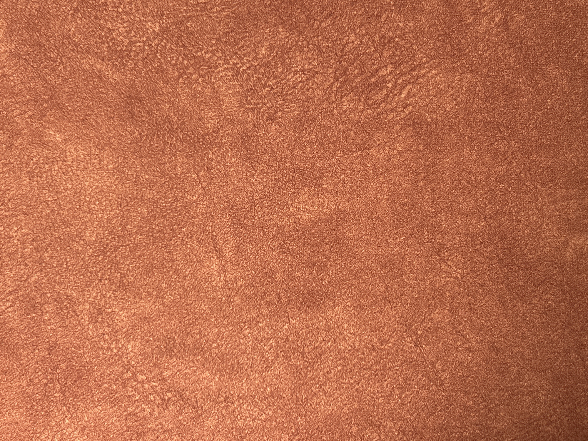 The media file presents an abstract, close-up view of a textured surface. The dominant color is a warm reddish-brown, resembling hues of copper, terracotta, or russet. The texture is finely granular and appears soft, suggesting materials such as suede, felt, or a brushed fabric. The surface is not uniformly colored; it displays subtle variations in shade with lighter and darker areas intermingling, creating a mottled, organic pattern and a sense of depth. These variations are consistent across the entire image, highlighting the material's tactile quality. There are no identifiable objects, people, or discernible activities taking place. No text is visible in the image.
