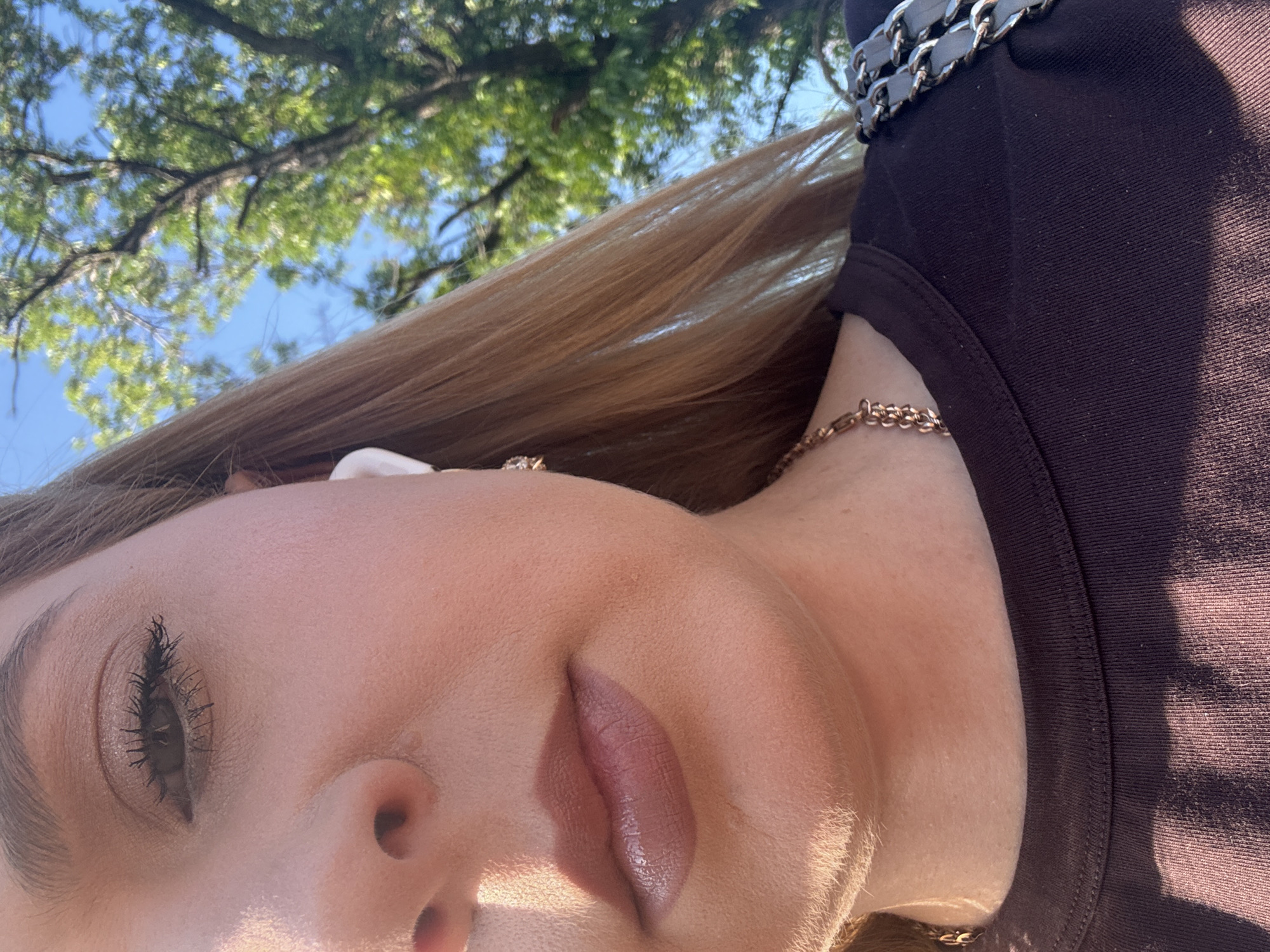 A half-faced, close-up portrait shows a fair-skinned woman from the nose down, with a close-up of her lips and eye. She is wearing a dark brown crew-neck t-shirt and a gold chain necklace. She has on light brown lipstick and mascara. A white wireless earbud is visible in her right ear. She has blonde hair. The background is out of focus, showing green trees and a blue sky. The lighting suggests it is daytime. The scene takes place outdoors in a park or a residential area with trees.