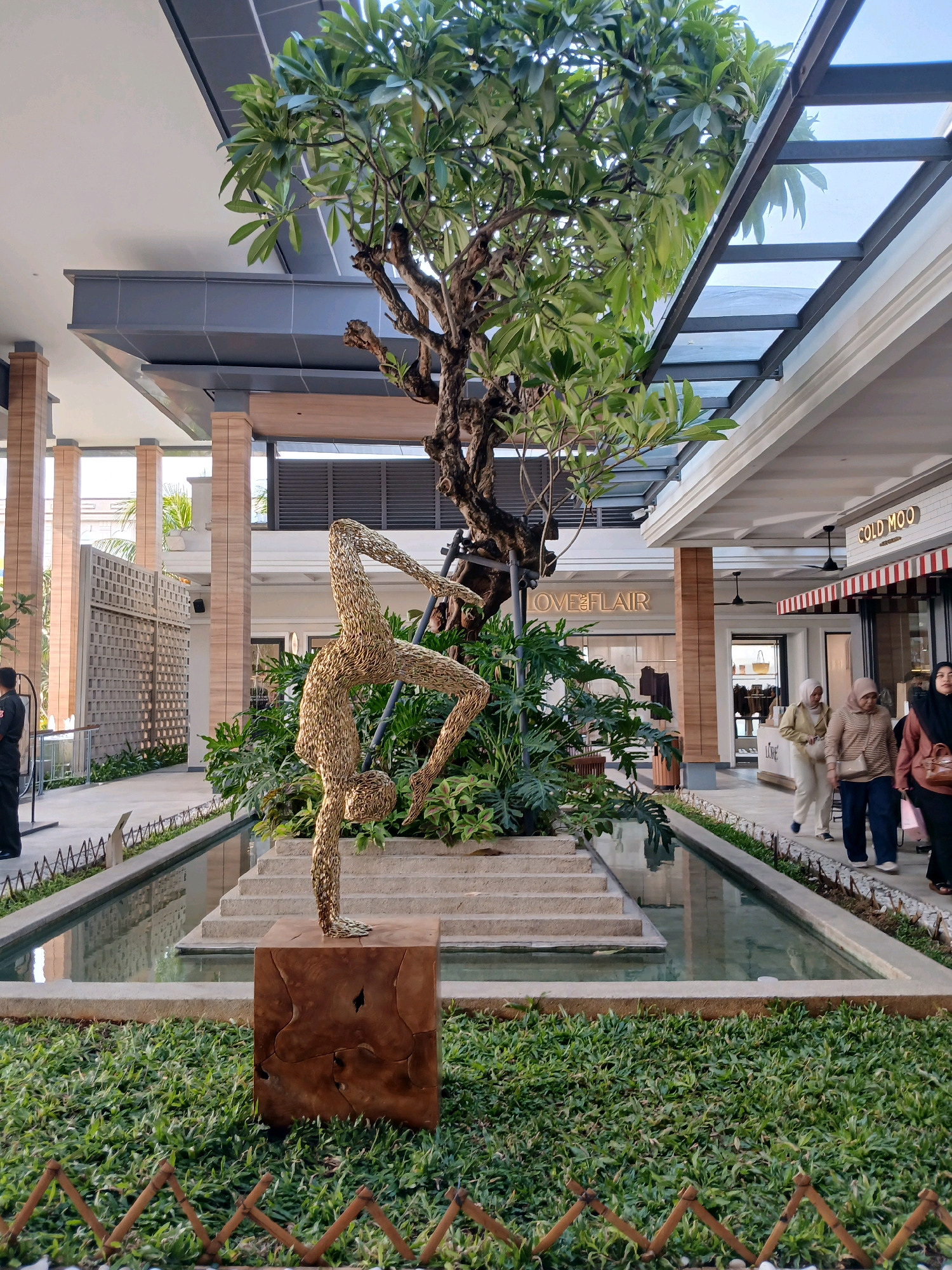 The image captures an outdoor area of a modern shopping complex in North Jakarta, Indonesia. In the foreground, a metallic sculpture resembling a gymnast in a contorted pose stands on a wooden pedestal, surrounded by lush green grass and a low, decorative fence. Behind the sculpture, a shallow water feature with stone steps leads to a small island with a large tree and dense foliage.
The background reveals the architecture of the shopping complex, with a glass roof and modern building structures. Several shops are visible, including one with a sign that reads "LOVE & FLAIR" and another with "COLD MOO". People are seen walking along the pathways of the complex. On the left side of the image, a security guard in a dark uniform stands by a railing. To the right, a group of women, some wearing hijabs, are walking past the shops. The scene is illuminated by natural light, suggesting it is daytime. The overall impression is of a well-maintained and aesthetically pleasing public space within an urban environment.