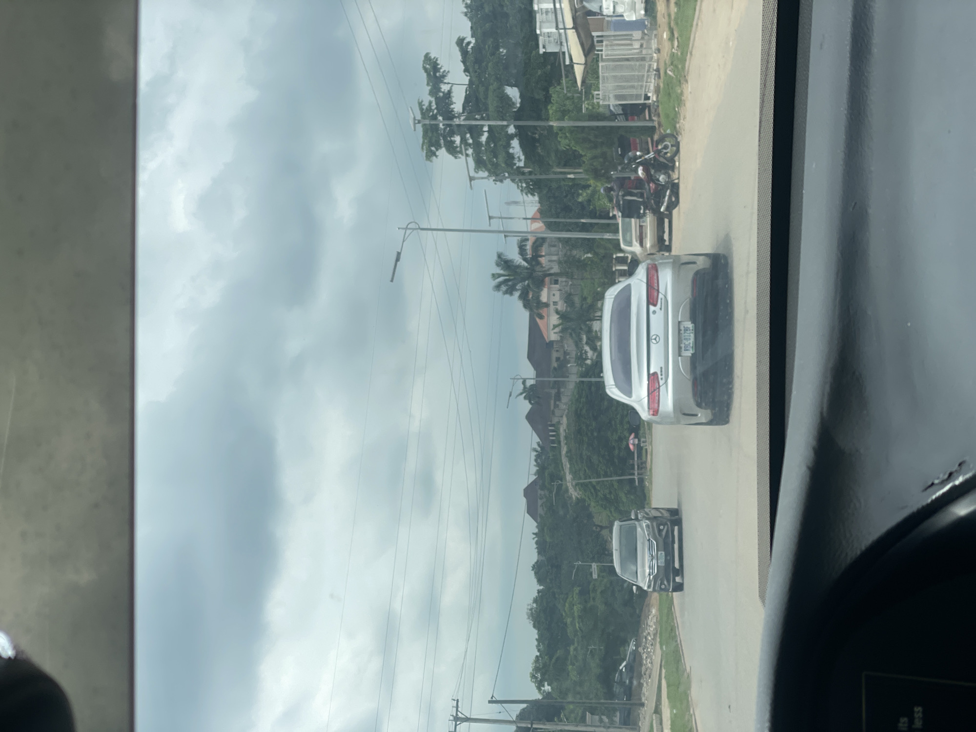 This is a forward-facing, eye-level, medium shot taken from inside a vehicle, looking out onto a street in Guzape, Nigeria. The sky is overcast with heavy clouds. The road is paved and appears to be a residential or commercial area.
In the foreground, the dashboard of the vehicle is visible. The main focus of the image is the street scene ahead. A silver Mercedes-Benz coupe is in the center of the frame, facing away from the camera. Its license plate is visible and reads "WBG 919F". To the left of the Mercedes, a gray minivan is traveling in the same direction. Further down the road, to the right, a red motorcycle with a rider wearing a black jacket and helmet is parked or stopped. Behind the motorcycle, a tan sedan is visible.
On the sides of the road, there are trees and various buildings. To the left, there is a utility pole with multiple wires. In the background, there are houses with distinctive rooflines, some of which appear to be large residential structures. There are also streetlights along the road. The overall environment suggests an urban or suburban setting with a mix of residential and possibly commercial properties.