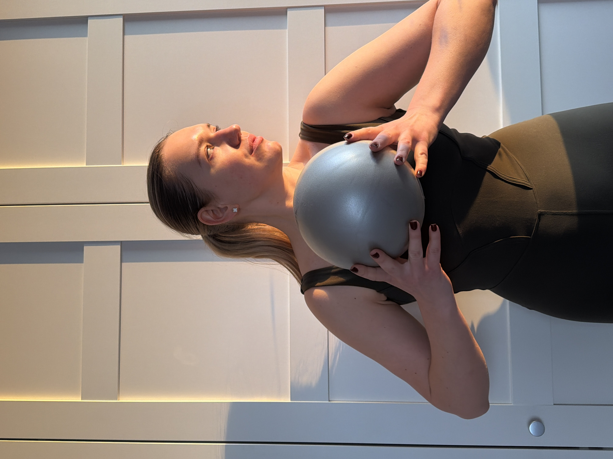A fair-skinned woman in an olive green sports bra and leggings is lying on her back, holding a gray exercise ball against her chest with both hands. She is facing upwards and slightly to her left. Her blond hair is pulled back into a low ponytail. The background consists of a white paneled wall with horizontal lighting. The location is Dreiliņi, Latvia.