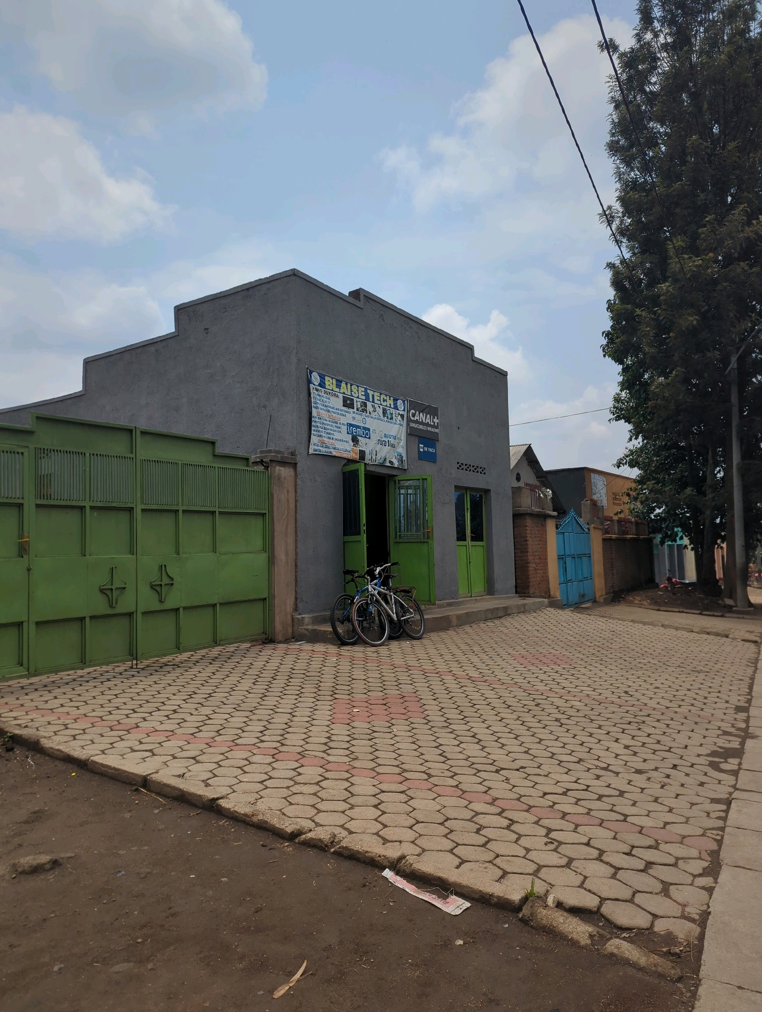The image shows a commercial building with a gray facade in Musanze District, Rwanda. The building has a large green metal gate to the left, with a honeycomb pattern in the center. In front of the building, a paved walkway with hexagonal tiles is visible, transitioning to a dirt road. Two bicycles are parked near the entrance of the building, which has green doors. A sign on the building reads "BLAISE TECH" and "CANAL+". The sky is partly cloudy. To the right of the building, a tall tree stands, and further down the street, other buildings and a blue gate are visible. The scene appears to be a street view of a business in a commercial area.
