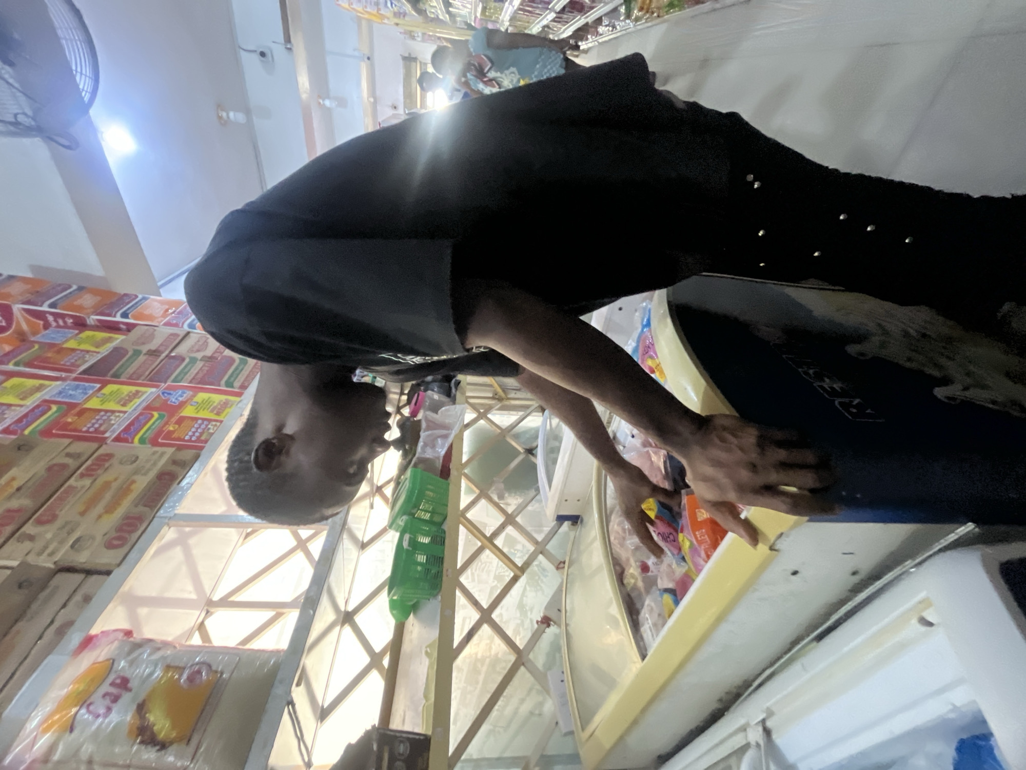 In Ado, Nigeria, a young man in a black t-shirt and black pants with silver studs is standing in a supermarket. He is leaning over a refrigerated display case filled with frozen goods, his hands reaching in to select items. Behind him, shelves are stocked with various products, including packaged noodles and other food items. Further in the background, another person, possibly a child, is visible near a shelf. The environment appears to be a typical retail setting with fluorescent lighting and display shelves.