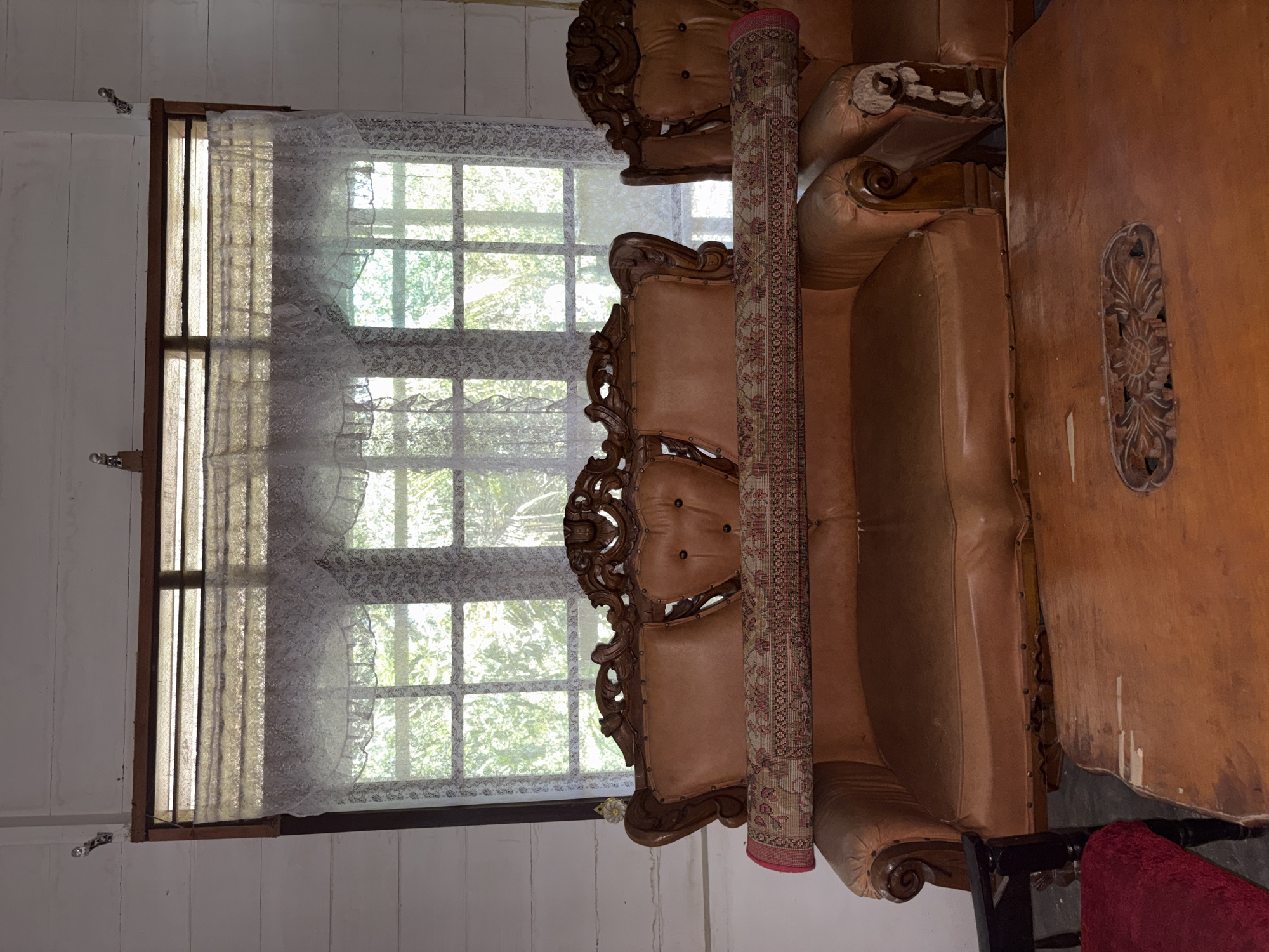 This image depicts the interior of a room, likely a living room, in a house located in Beunyot, Indonesia. Dominating the center of the frame are two adjacent brown leather sofas with ornate wooden trim. A rolled-up patterned rug is placed across the backs of the sofas. Behind the sofas is a large window covered by white lace curtains, through which greenery is visible, suggesting an outdoor environment. The walls are paneled with white wood. In the foreground, a wooden coffee table with carved details is partially visible. The overall impression is of a traditional, possibly older, domestic setting. There are no people present in the image.