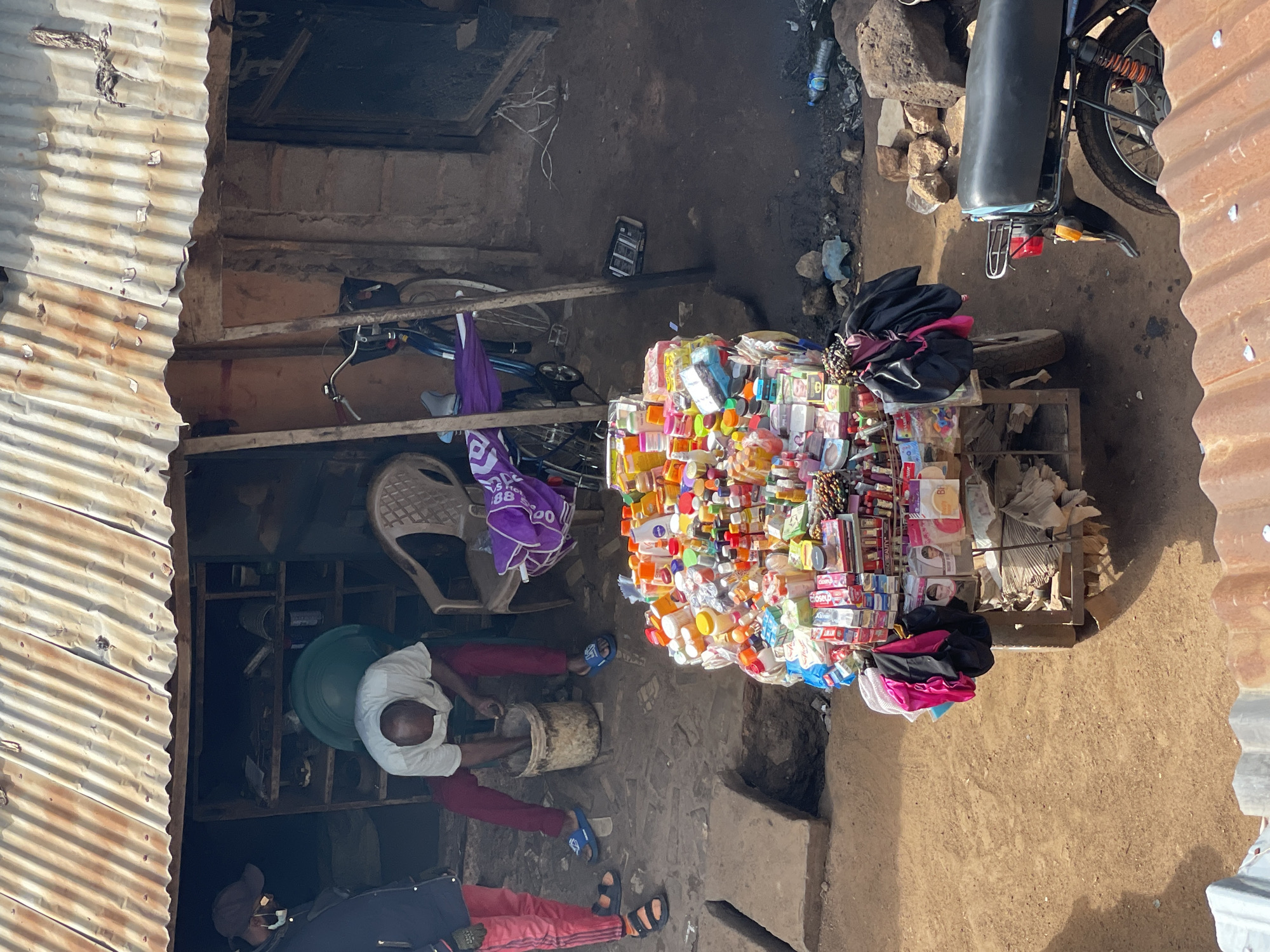 In an outdoor marketplace in an unknown city, a man wearing a white shirt and red pants is bent over a bucket in front of a corrugated metal structure. To his left, a person in a dark jacket and red pants stands with their back to the camera. In the foreground, a cart is laden with various products, including bottles, boxes, and packets of toiletries and cosmetics, some of which are arranged in colorful displays. Behind the cart, a motorcycle is parked. The ground is dirt and debris, and the overall environment appears rustic and informal.