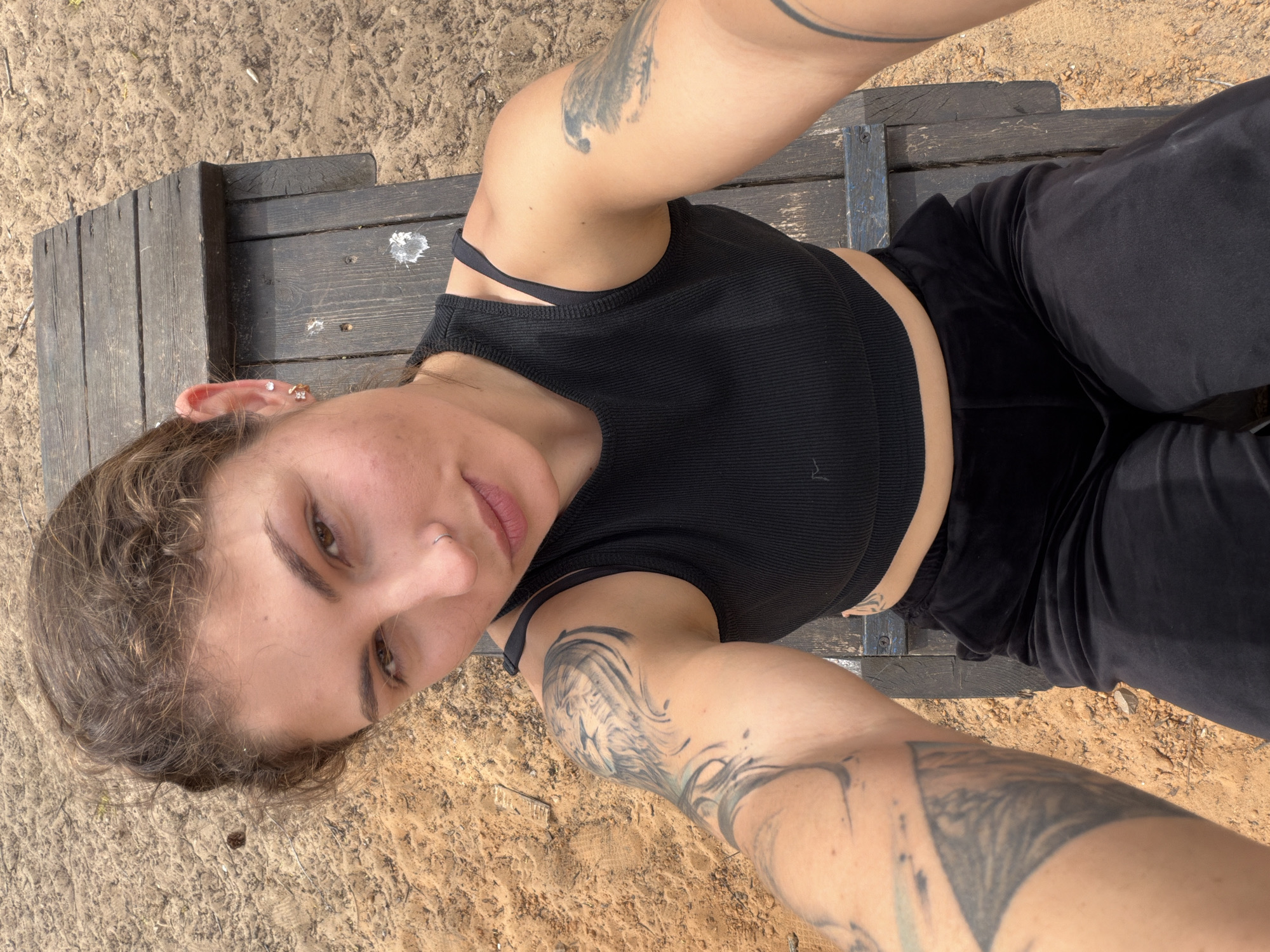 A person with arm tattoos lies on a wooden surface in an outdoor setting, possibly in Kiryat Bialik, Israel. The person wears a black top and black pants. The surface beneath is sandy.