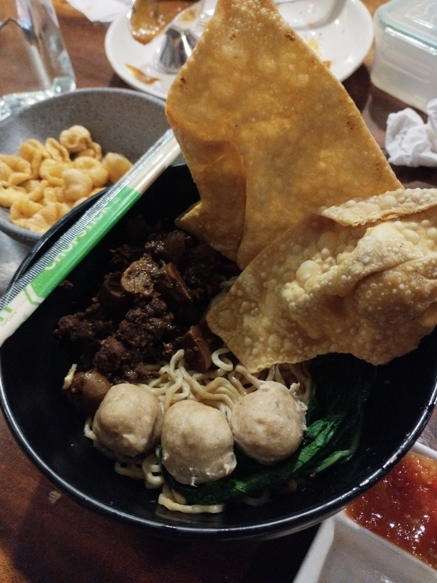 The image shows a close-up of a bowl of noodles with meatballs and fried wontons. A pair of chopsticks with a green wrapper rests on top of the noodles. To the left of the bowl, a small grey bowl contains some fried snacks. In the foreground, a small white dish holds a red sauce, likely chili sauce. The setting appears to be a restaurant in the city of Pematang Siantar, Indonesia, with wooden furniture visible in the background. The lighting is warm and diffused, suggesting an indoor environment. There are no people visible in the image.
