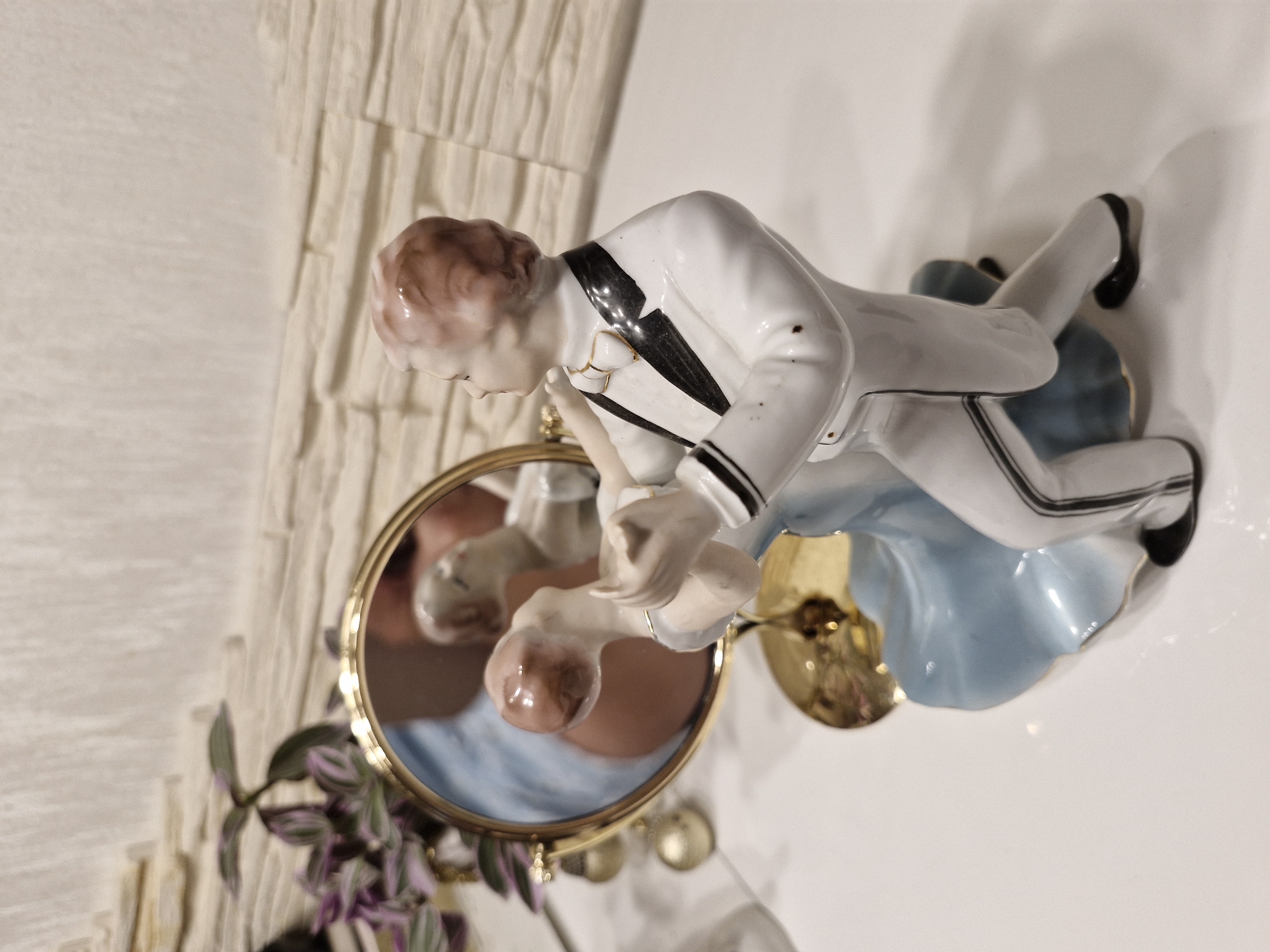 A close-up, eye-level shot shows a porcelain figurine of a couple dancing, positioned on a white surface. The male figure wears a white tuxedo with black lapels and trim, and a white bow tie. The female figure is adorned in a flowing light blue gown. They are reflected in a golden-rimmed mirror that stands behind them. A plant with purple and green leaves and several golden baubles are visible to the left of the mirror. The background features a textured, off-white wall. The scene is set indoors, on a white tabletop, with no discernible connection to the city of Bremen, Germany.