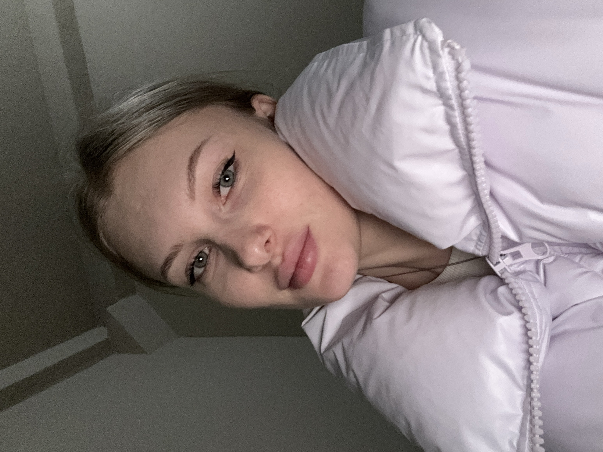 A headshot of a young adult female is captured indoors under low, artificial lighting. The subject, positioned centrally and looking directly at the viewer, has fair skin, blonde hair, and blue-green eyes with prominent black winged eyeliner. Her lips appear augmented, and a subtle smile is present. She is dressed in a light purple, possibly lavender, puffer jacket featuring a high, padded collar that partially obscures her lower face on the right side. The jacket is zipped approximately halfway, revealing a light-colored undergarment and a thin, dark necklace. The background consists of dark, indistinct surfaces, with subtle geometric lines visible in the upper left, indicating interior architectural features. The precise location within the city of Perm, Russia, is not ascertainable from the visual elements depicted.