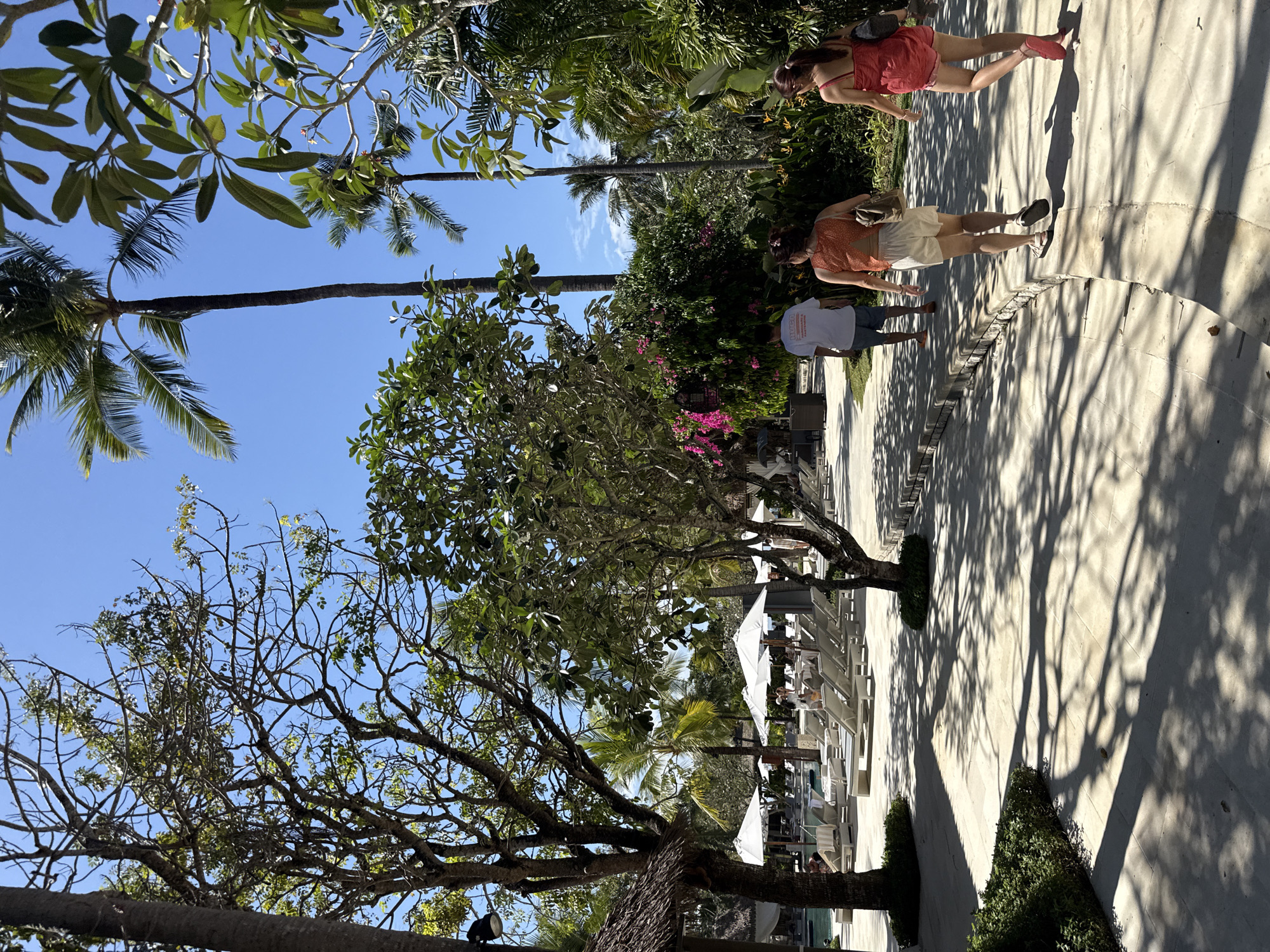 In Kuta Selatan, Indonesia, this outdoor scene features a paved walkway lined with trees, casting dappled shadows. Several palm trees and other tropical foliage are visible, with a bright blue sky overhead. In the background, lounge chairs and white umbrellas suggest a resort or hotel pool area. Three individuals are walking away from the camera on the walkway. One person, wearing a white t-shirt and blue shorts, is furthest ahead. Following behind is a person in an orange top and white shorts, and closest to the camera is a person in a red dress. The overall atmosphere is sunny and tropical.