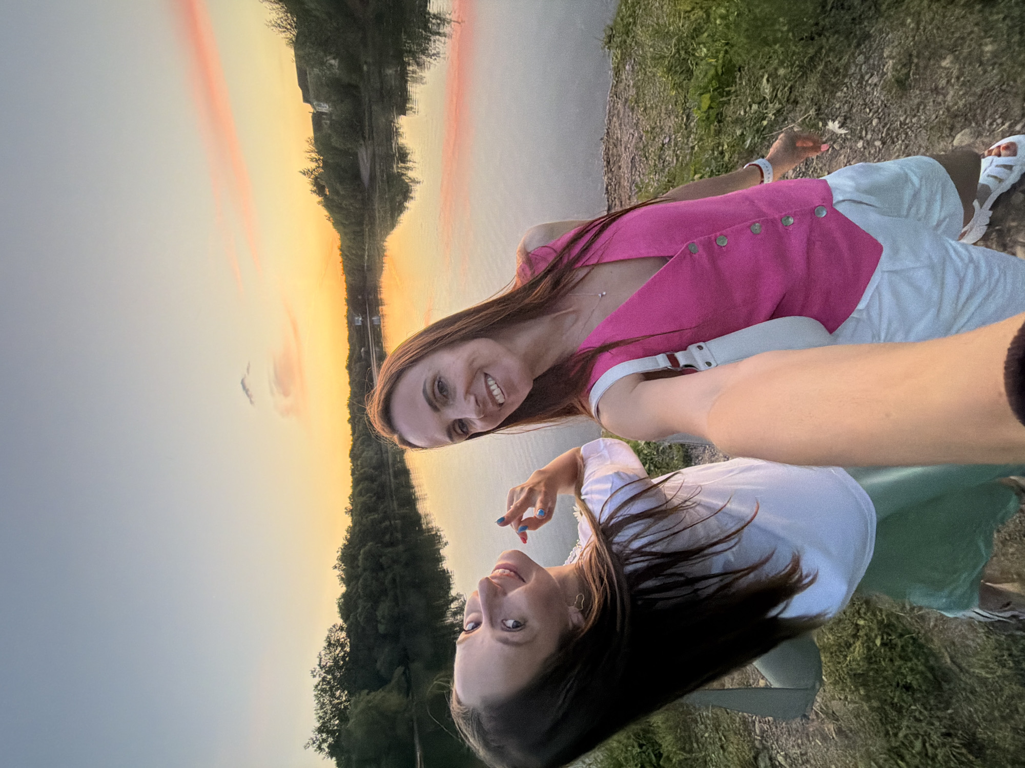 Two women are taking a selfie at sunset next to a body of water. The woman on the left is wearing a white t-shirt and a green skirt, and she is looking up and to the left with a wide smile. The woman on the right is wearing a pink vest and white shorts, and she is looking at the camera with a smile. The water is calm and reflects the colors of the sunset, which is a mix of orange, yellow, and pink. Trees line the opposite bank of the water. The ground in the foreground is covered with dirt and grass. The image appears to have been taken outdoors in a natural setting, possibly a park or a nature reserve. The overall mood is happy and relaxed.