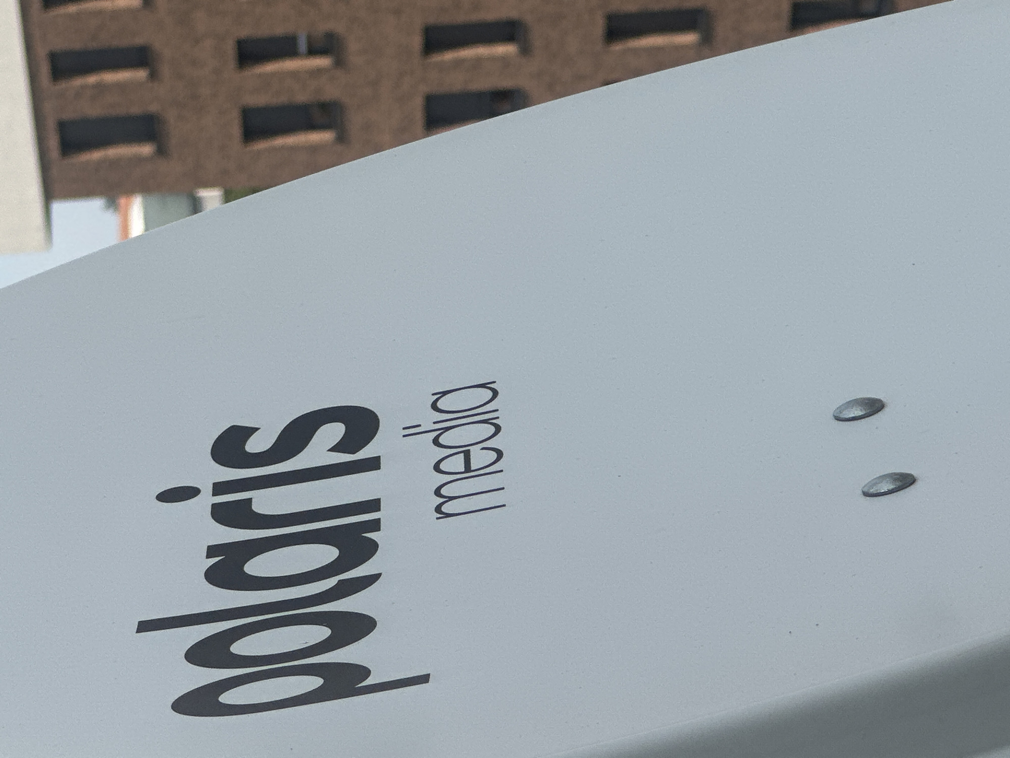 The image shows a white, curved surface with the black text "polaris" in a stylized font and "media" underneath it. The background is blurred, showing a brown building with multiple windows, suggesting an urban environment. The lighting suggests it is daytime. There are two metallic rivets on the white surface. The image appears to be a close-up of a sign or a piece of equipment. There is no information available about the location in Belgrade, Serbia.