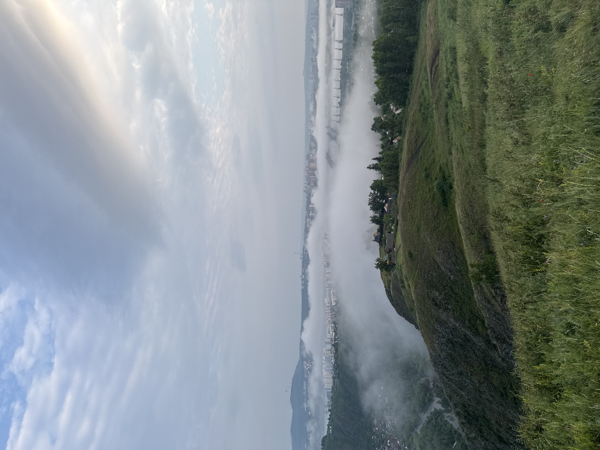 The image captures a panoramic view of Krasnoyarsk, Russia, dominated by a thick layer of fog that blankets the city. The foreground features a grassy slope dotted with scattered vegetation and a few small houses. A steep, rocky incline slopes down to the left, revealing a small river winding through a densely forested valley.
The midground is characterized by a sprawling cityscape partially obscured by fog. Numerous apartment buildings, ranging in height and color, are visible, their structures appearing softened and blurred by the atmospheric conditions. Industrial elements, such as cranes and bridges, emerge from the mist, suggesting a port or industrial zone along the river.
In the background, rolling hills and mountains form the distant horizon, their outlines indistinct against the overcast sky. The sky itself is a mix of pale blue and soft grey clouds, with patches of sunlight breaking through on the right, casting a gentle glow over the scene. The overall impression is one of a serene, misty morning over a vast urban landscape, with nature and industry coexisting under a hazy sky. There are no visible people or animals in the frame.