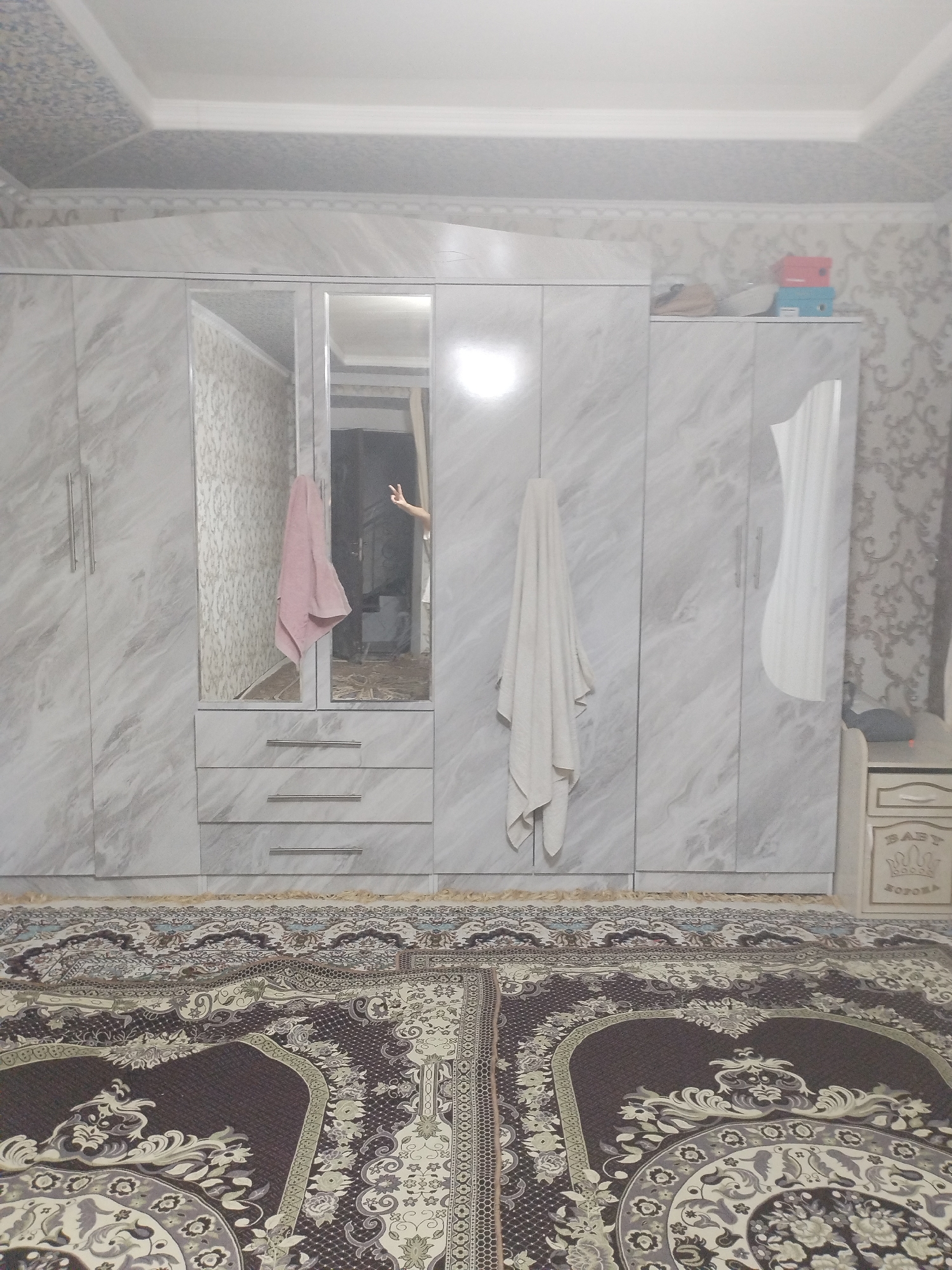 A bedroom scene in Kyrgyzstan, during daytime. The room is furnished with a large, modern wardrobe with a marble-effect finish. The wardrobe has multiple doors and drawers, and features a mirrored section. Two towels are visible, one pink and one white, hanging from the wardrobe. A person's hand is visible in the mirror, giving a peace sign. On top of the wardrobe, two colorful boxes are stacked. To the right of the wardrobe, a small nightstand with decorative carvings is present. The floor is covered with a patterned rug. The ceiling is decorated with patterned panels and molding. The overall impression is of a tidy and organized room, likely a personal living space. The style of the furniture and decor suggests a contemporary aesthetic within a socialist context, common in Kyrgyzstan. There is no visible text in the image.