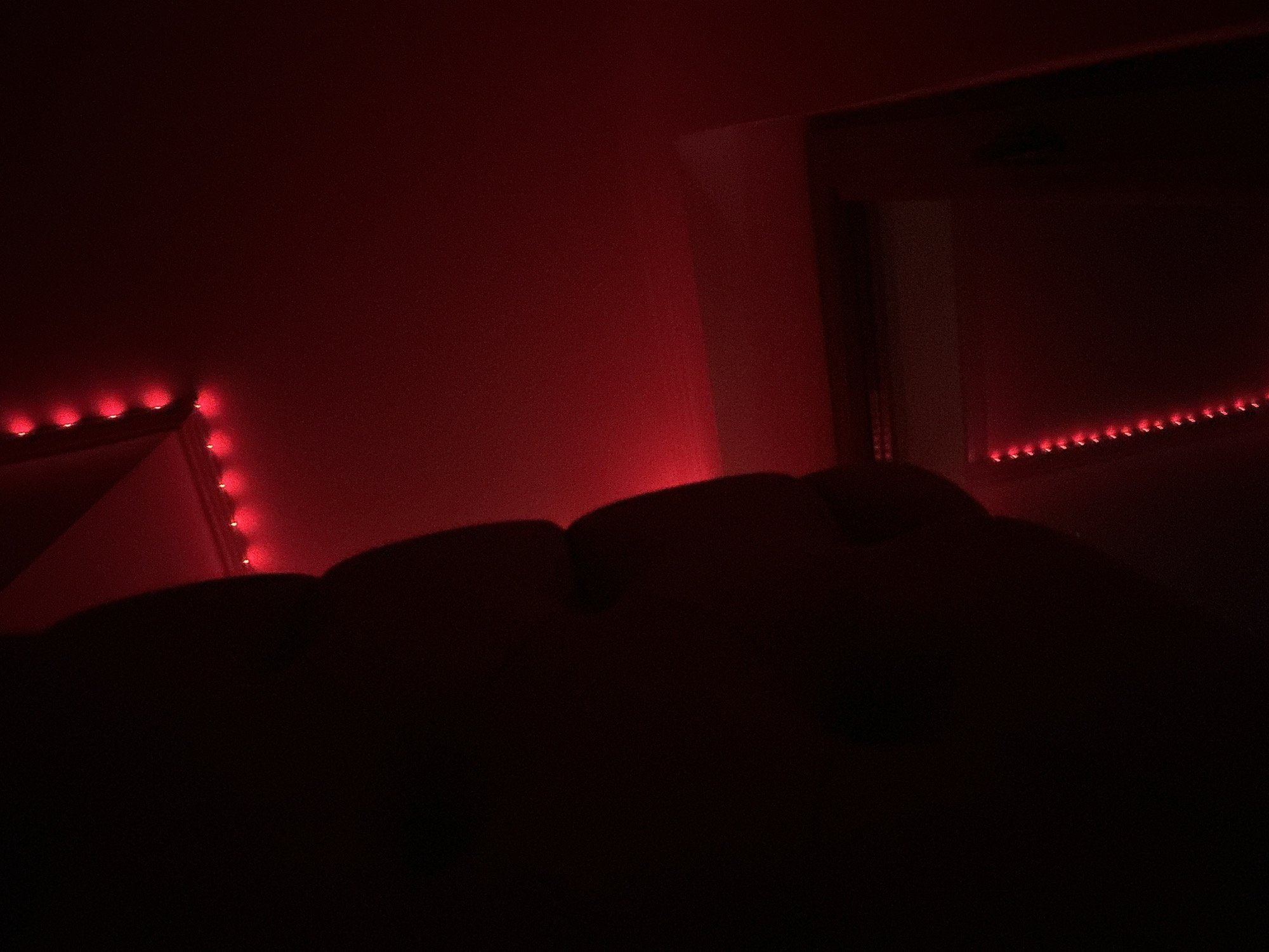 The image depicts a dimly lit room illuminated by red LED strip lights. In the foreground, a dark, amorphous shape, possibly furniture or a person covered in shadow, occupies the lower half of the frame. The background features a wall bathed in a strong red light, creating a gradient effect that fades into darkness. Red LED lights are visible along the top edge of a corner in the left background and in a strip along the top of a wall or surface in the right background. The overall impression is of a room set for ambiance, with the red lighting casting a dramatic and somewhat abstract mood. There is no indication of the city of Shumen, Bulgaria within the image itself.