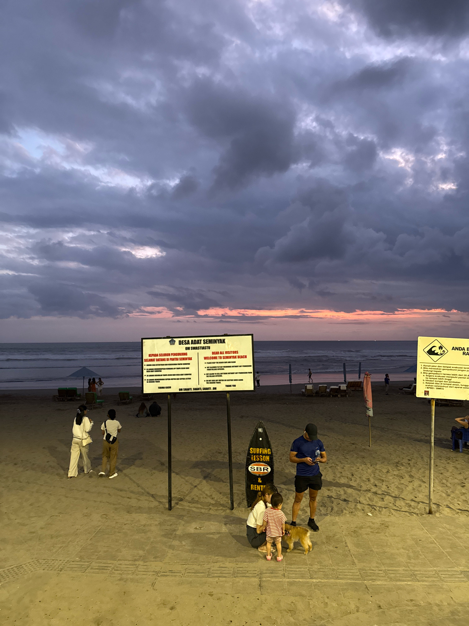 An overcast sunset over the ocean at Seminyak Beach in Kuta, Indonesia, is depicted. The sky is filled with dramatic, dark clouds tinged with hues of purple and pink from the setting sun. The ocean waves are visible, and the beach is made of sand.
In the foreground, two people are standing near a sign that says "DESA ADAT SEMINYAK OM SWASTIASTU". Another sign, also in Indonesian and English, is visible to the right. In the middle ground, a family with a small dog is interacting. A man in a blue shirt and black shorts is looking at his phone, while a woman in white is crouching down, attending to a child in a striped shirt and pink shoes. The dog is a light brown color.
Further back on the beach, several lounge chairs and umbrellas are set up. A few other people are scattered across the sand, some walking and others sitting. A black surfboard with "SURFING LESSON & RENT" written on it stands upright near the family and dog. The overall atmosphere appears calm, despite the dramatic sky.
