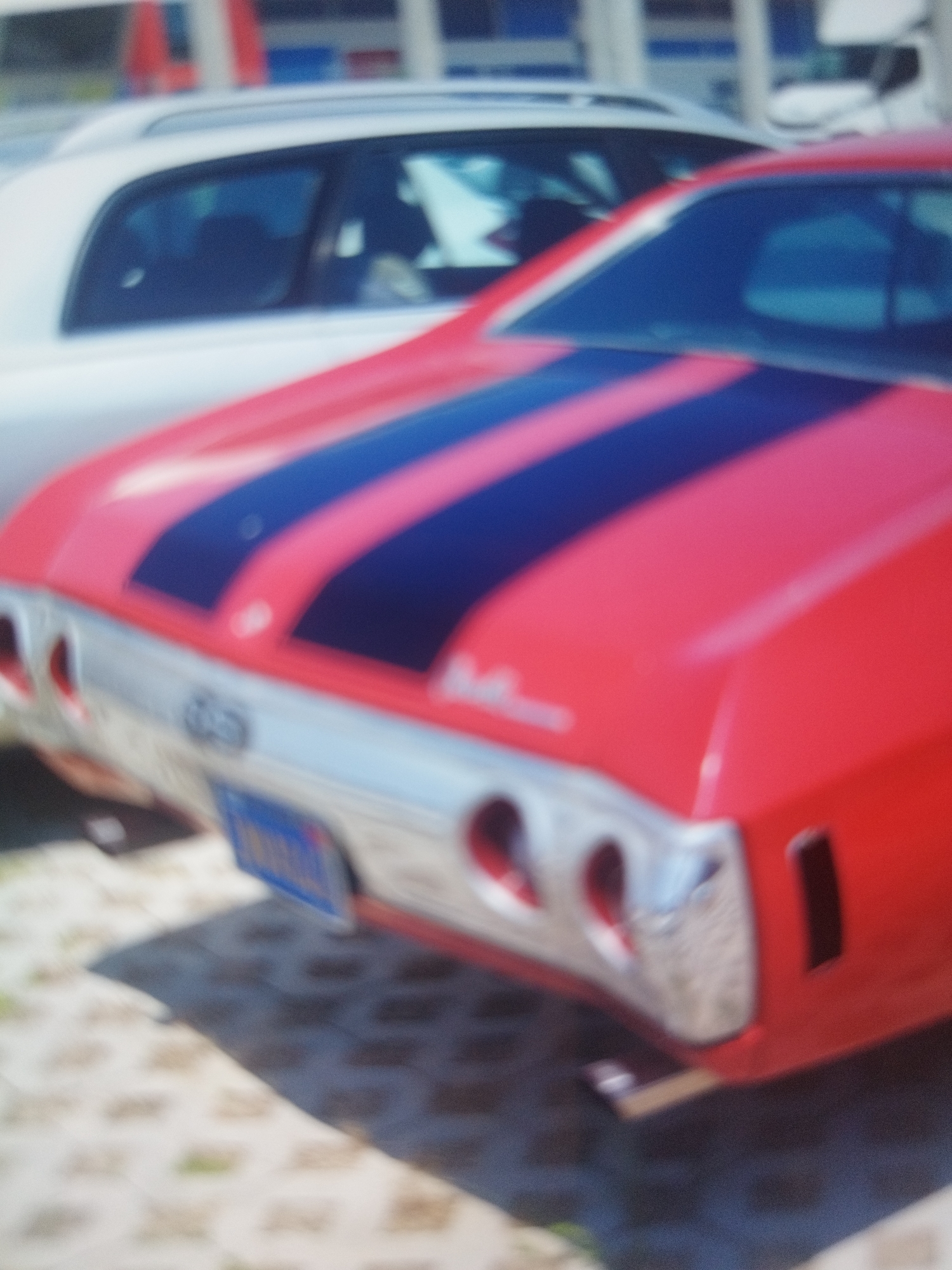 The image depicts the rear of a red classic car with black racing stripes parked on a patterned surface. The car has chrome detailing and a blue license plate. Behind the red car is a white car. The location is not discernible from the image.