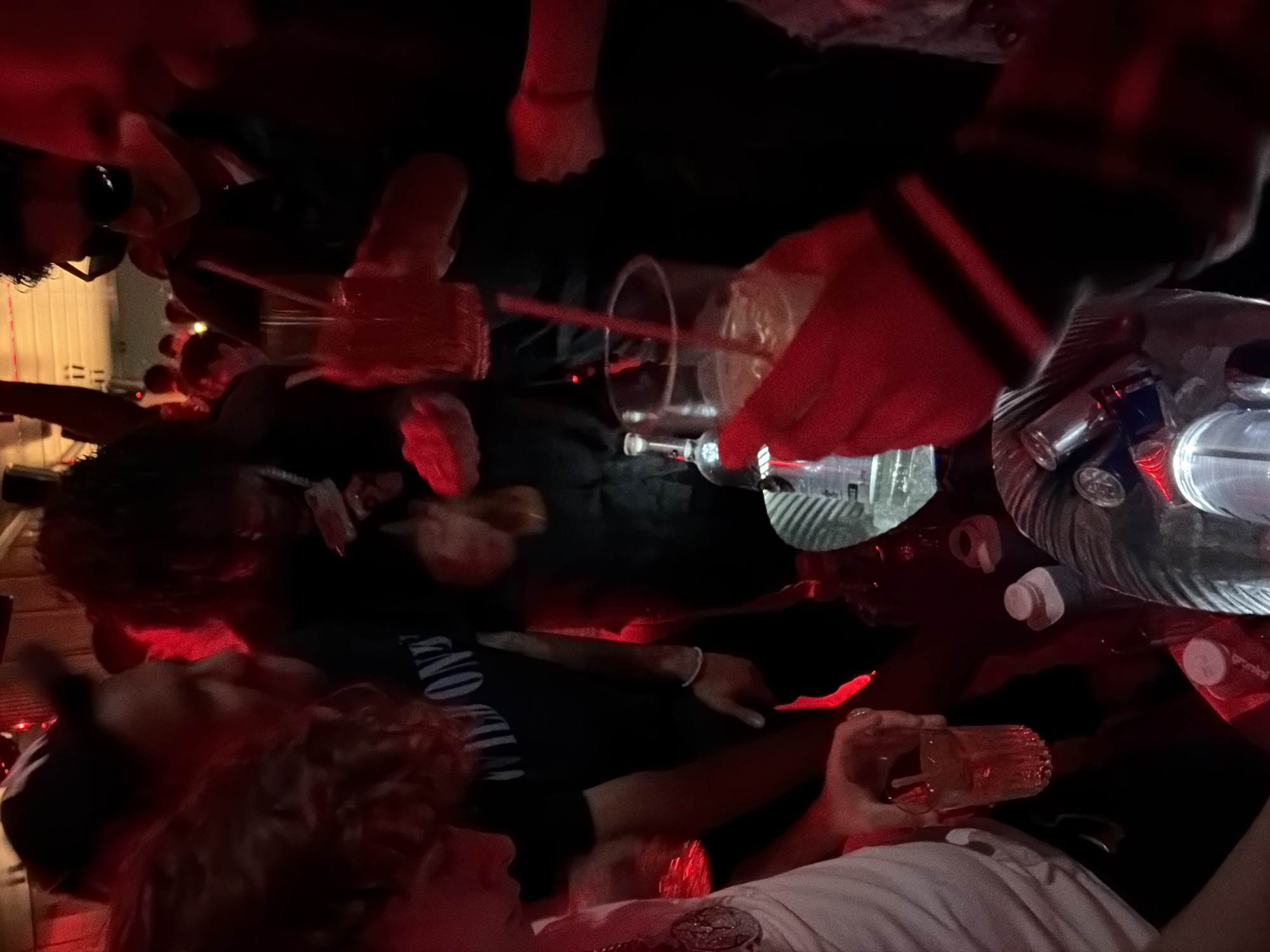 The scene captures an indoor social gathering in a dimly lit environment, characteristic of a nightclub or lounge, located in Paris, France. The pervasive red ambient lighting illuminates multiple individuals clustered around a table. A central figure with dark, curly hair wears a black t-shirt with "WILD ONE" visible and holds a stack of Euro banknotes along with a small bottle. To the right, another individual wearing sunglasses holds a tall glass containing a red beverage with a straw. On the left, a person in a white t-shirt with a large medallion holds a clear glass with a red drink. Prominently displayed on the table is a clear ice bucket containing bottles, including a transparent vodka bottle, amidst ice. Additional clear glasses with various beverages and straws are visible on the table and in the hands of other partially visible individuals, indicating active consumption and social interaction.