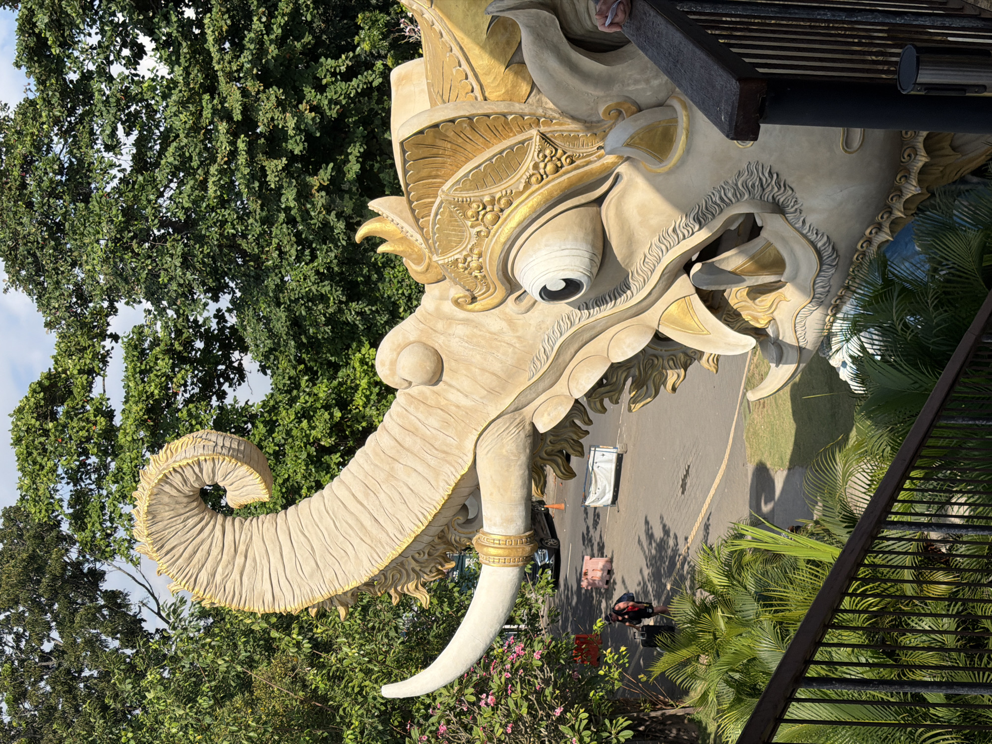 An outdoor, eye-level, medium shot captures a close-up of a large, ornate elephant statue in Denpasar, Indonesia. The statue, primarily beige with gold accents, dominates the frame with its detailed head, curled trunk, and large, staring eye. The statue appears to be part of a larger structure, possibly an entrance or landmark, with a dark wooden railing and metal posts visible in the foreground.
In the background, a paved area suggests a road or parking lot. A person, appearing to be a tourist with a backpack and rolling suitcase, walks away from the camera towards the left side of the frame. Other objects in the background include parked vehicles, construction barriers, and traffic cones, indicating ongoing work or an event. Lush green trees and foliage frame the scene, with a partly cloudy sky visible above. The sunlight casts shadows, suggesting a bright, sunny day.