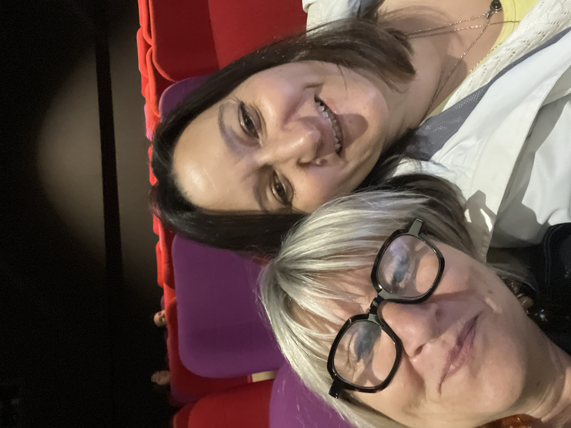 Two women are posing for a selfie in a seated area, likely a theater or auditorium, based on the red and purple upholstered seats visible in the background. The woman at the top of the frame has dark hair and is smiling. She is wearing a light-colored jacket and a yellow top with a necklace. The woman at the bottom of the frame has short, gray hair and is wearing black framed glasses. The dark background suggests the location is indoors. The city of Montbrison, France is not visibly represented in the image.